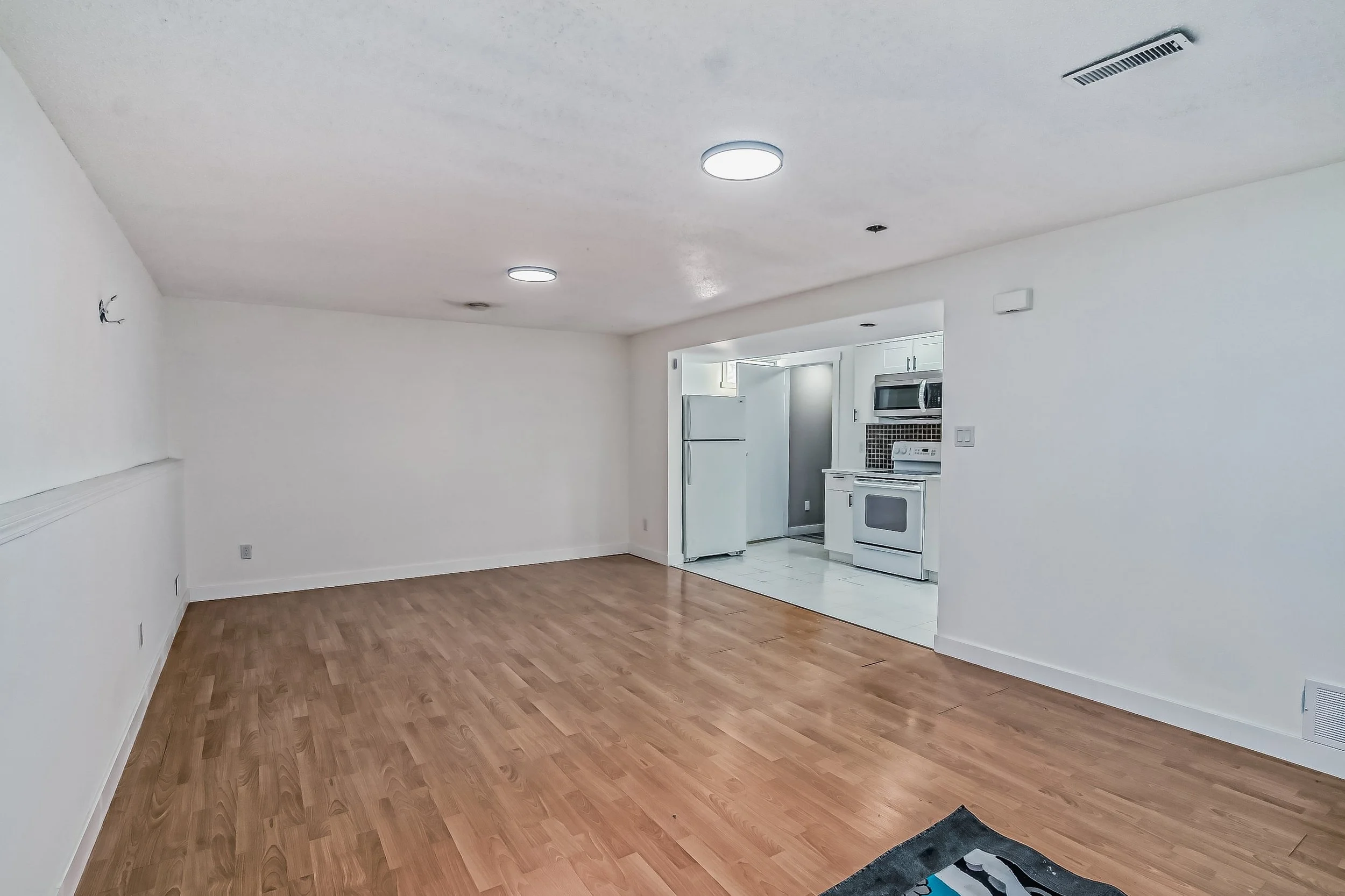 Empty living room with white walls and wood laminate flooring, open kitchen with white appliances and tiled floor.