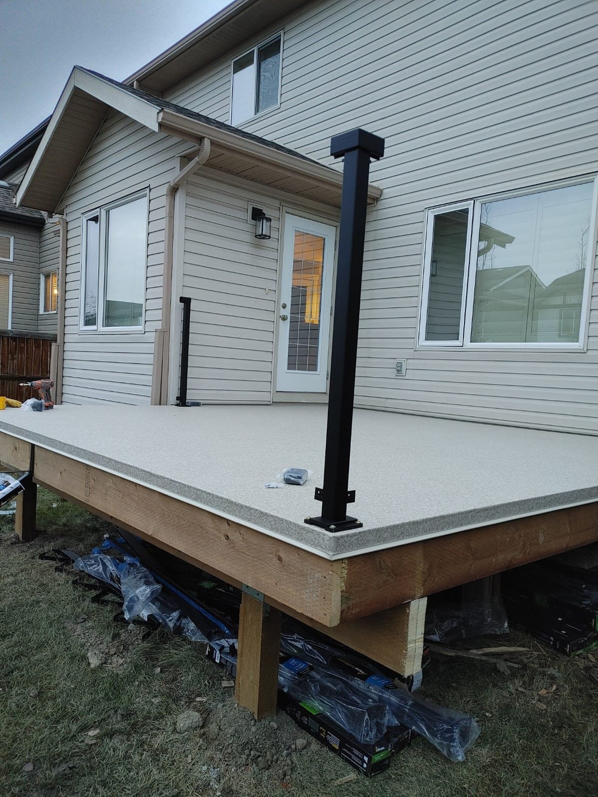 A newly built elevated backyard patio with beige decking, black metal railing posts, attached to a beige house with vinyl siding, multiple windows, and a door with glass panes. The patio's support structure is visible underneath, with construction to