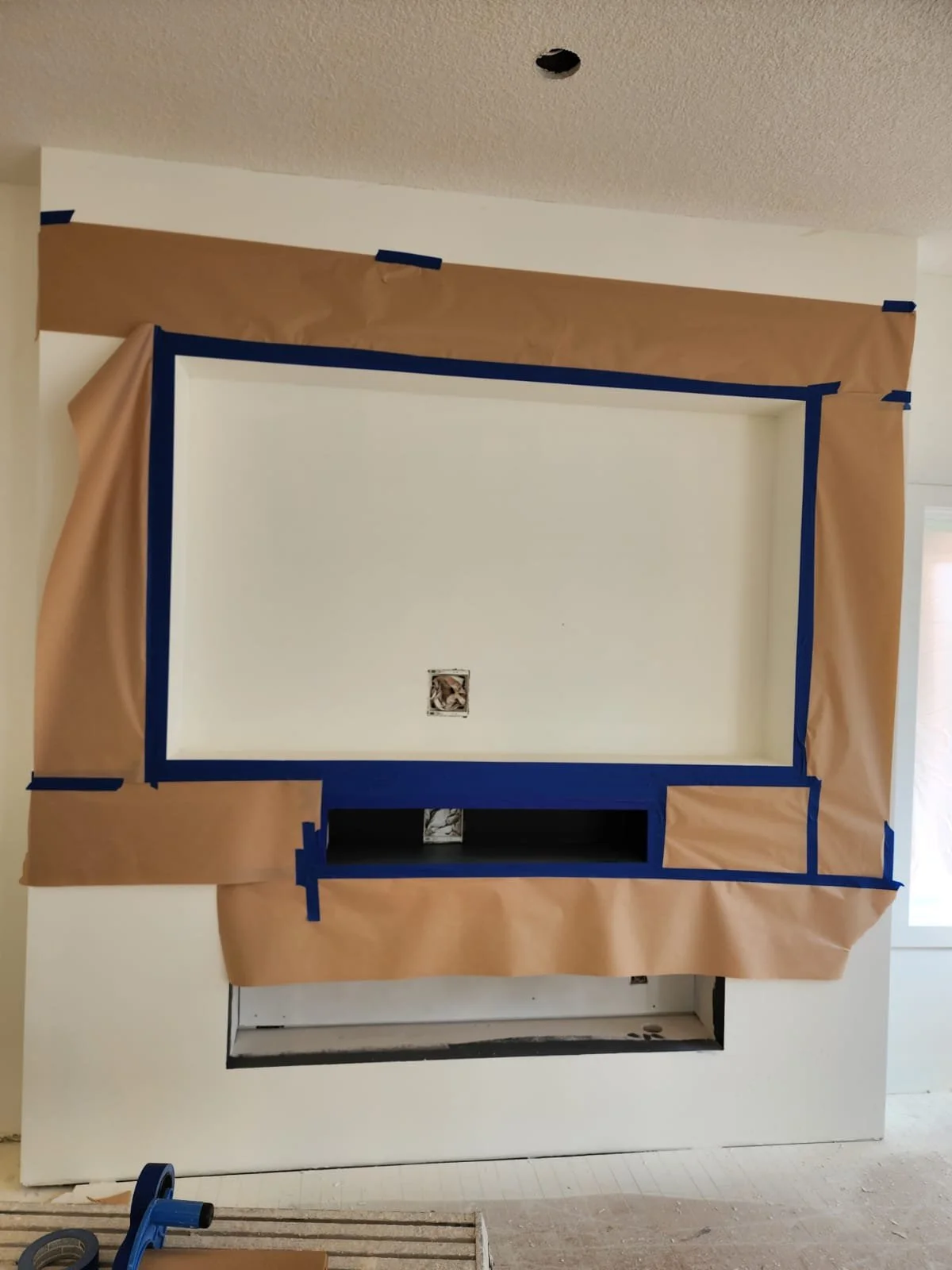 Wall with an unfinished fireplace, surrounded by masking paper and painter's tape for painting or renovation.