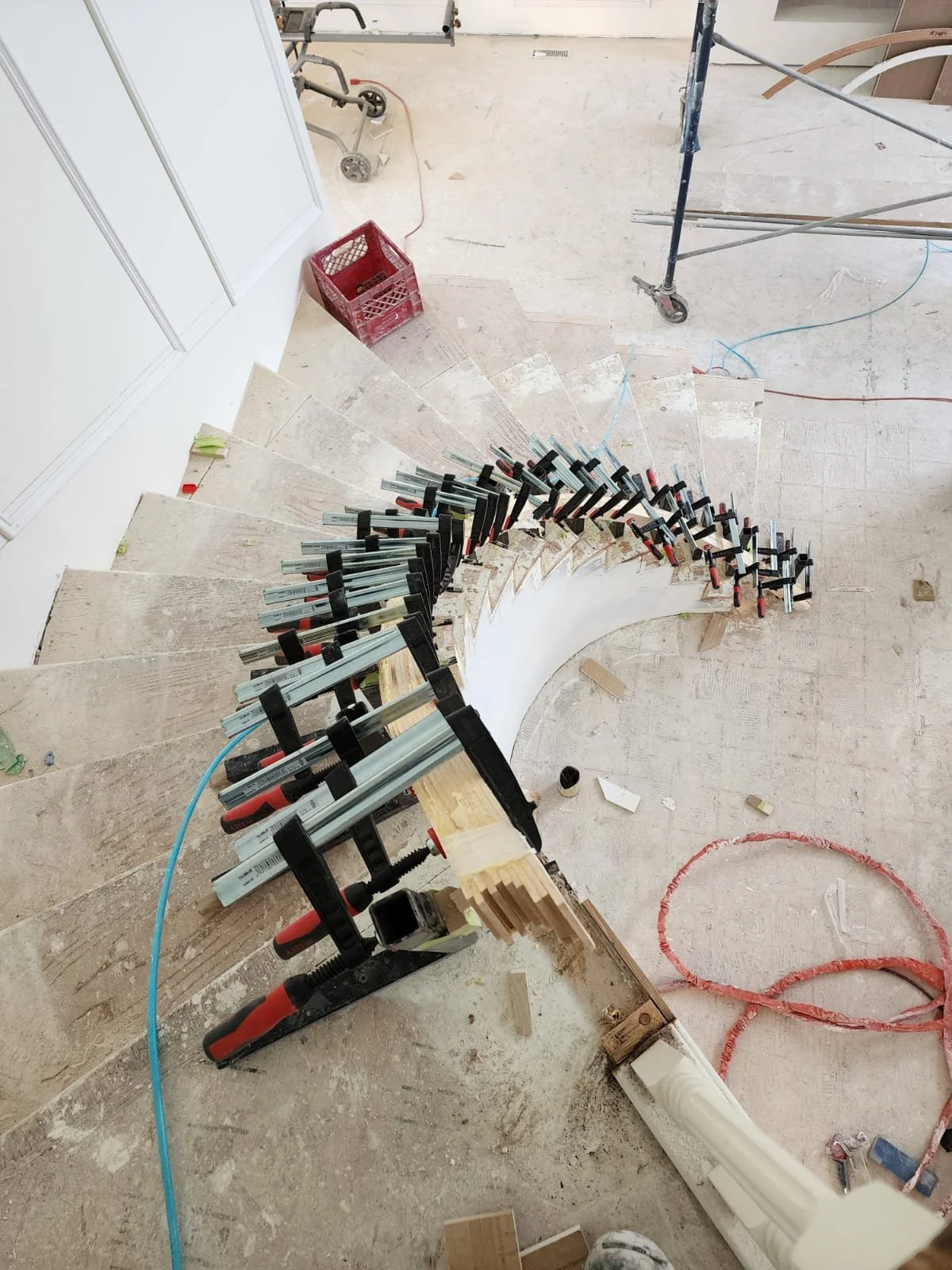 Construction site showing an unfinished curved staircase with multiple clamps holding temporary supports on the steps, and various tools and materials scattered around.