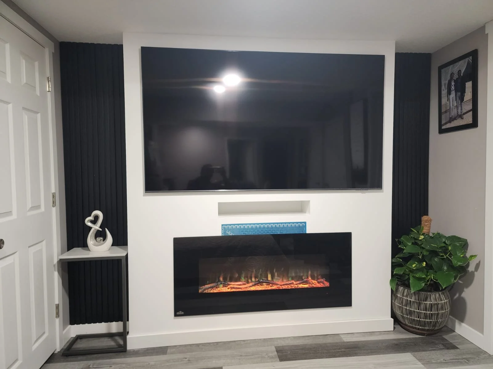 Living room with a large flat-screen TV mounted above a modern electric fireplace, black vertical panel accents on both sides, a potted plant on the right, a framed black-and-white photograph on the wall, and a small decorative swan sculpture on a stand to the left.