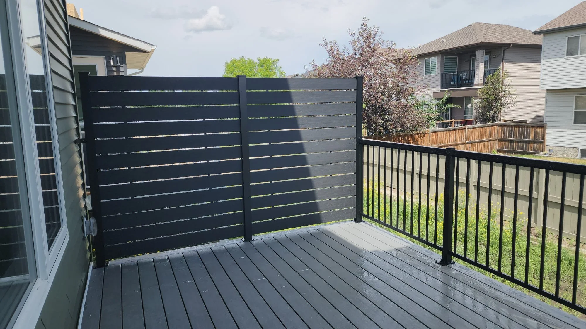 View of a residential backyard patio with black wooden privacy screen and black metal railing, with neighboring houses and trees in the background.