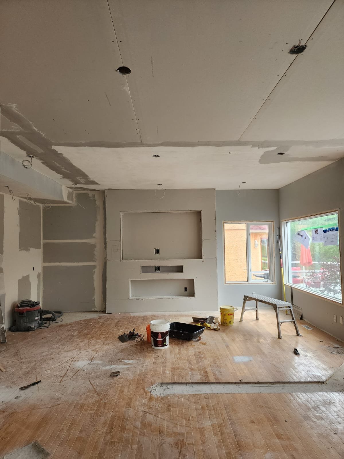 Interior of a room under renovation with unfinished drywall, tools, and construction materials scattered on the wooden floor. There are two large windows and an alcove in the wall, with exposed wiring and partially painted surfaces.