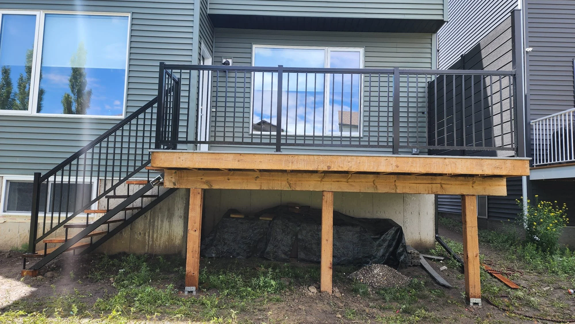 Back of a house with a new raised deck under construction, with black metal railing, wooden supports, and stairs on the left side, overlooking a yard with grass and some construction materials.