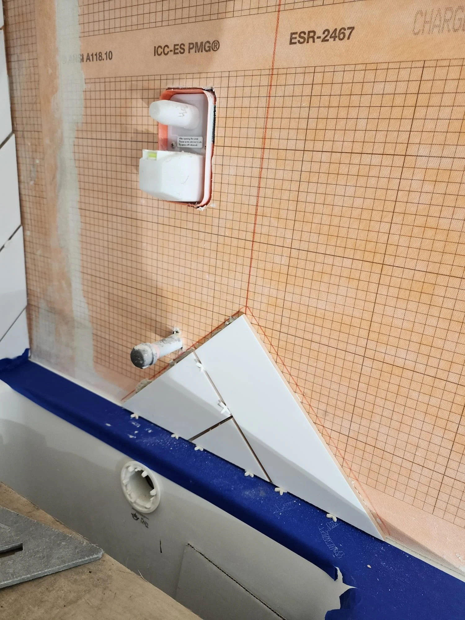 Close-up of a bathroom wall under construction with a exposed light switch or outlet, partially tiled wall, and a section of the bathtub or shower base with visible tiles and construction materials.