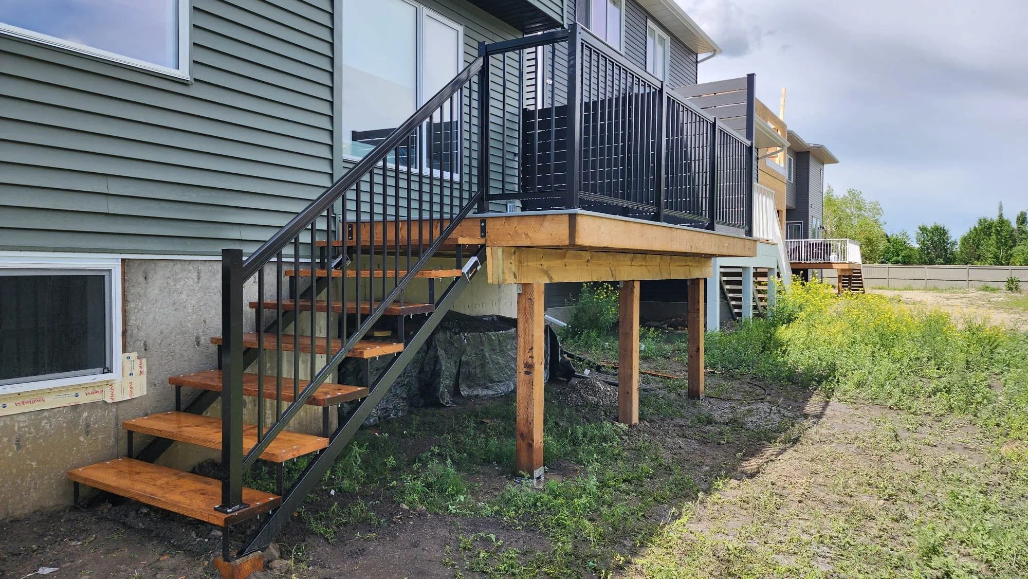 Newly built wooden deck with black metal railing attached to the back of a house, with stairs leading down to the yard. The house has gray siding and a concrete basement window, with some construction materials visible underneath.