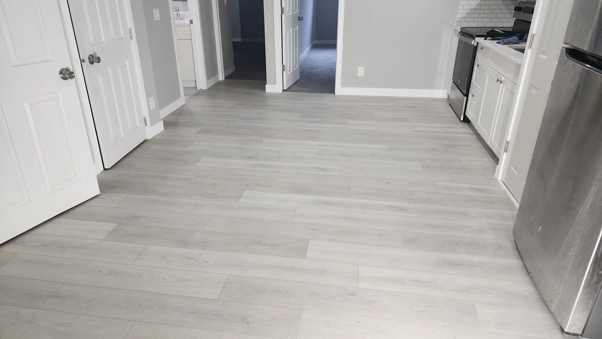 Empty kitchen with light gray wood flooring, white cabinets, stainless steel appliances, and an open doorway leading to a room with gray carpet.