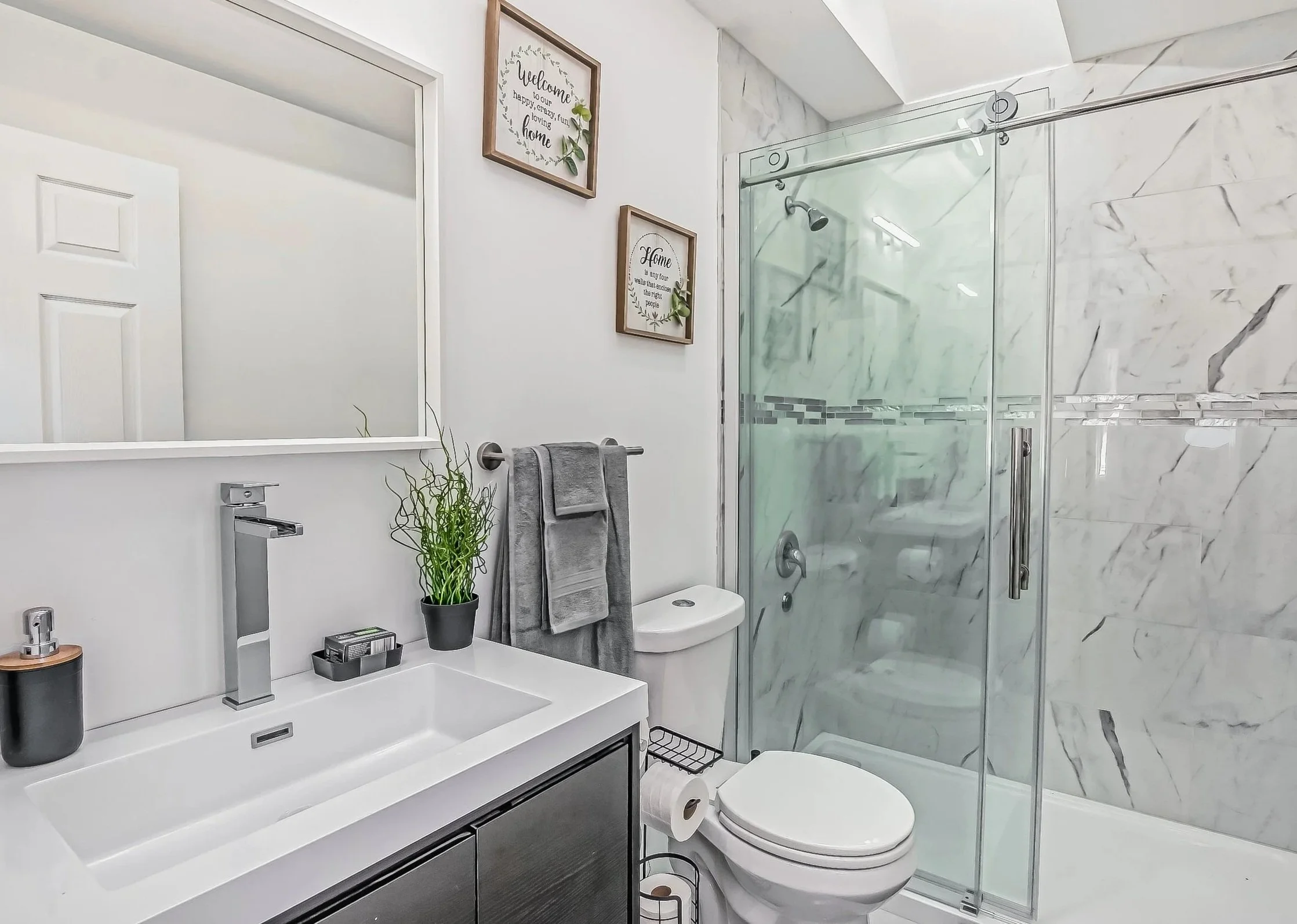 Modern bathroom with marble shower, white walls, gray towels, a mirror, and a vanity with a sink, plants, and decor.