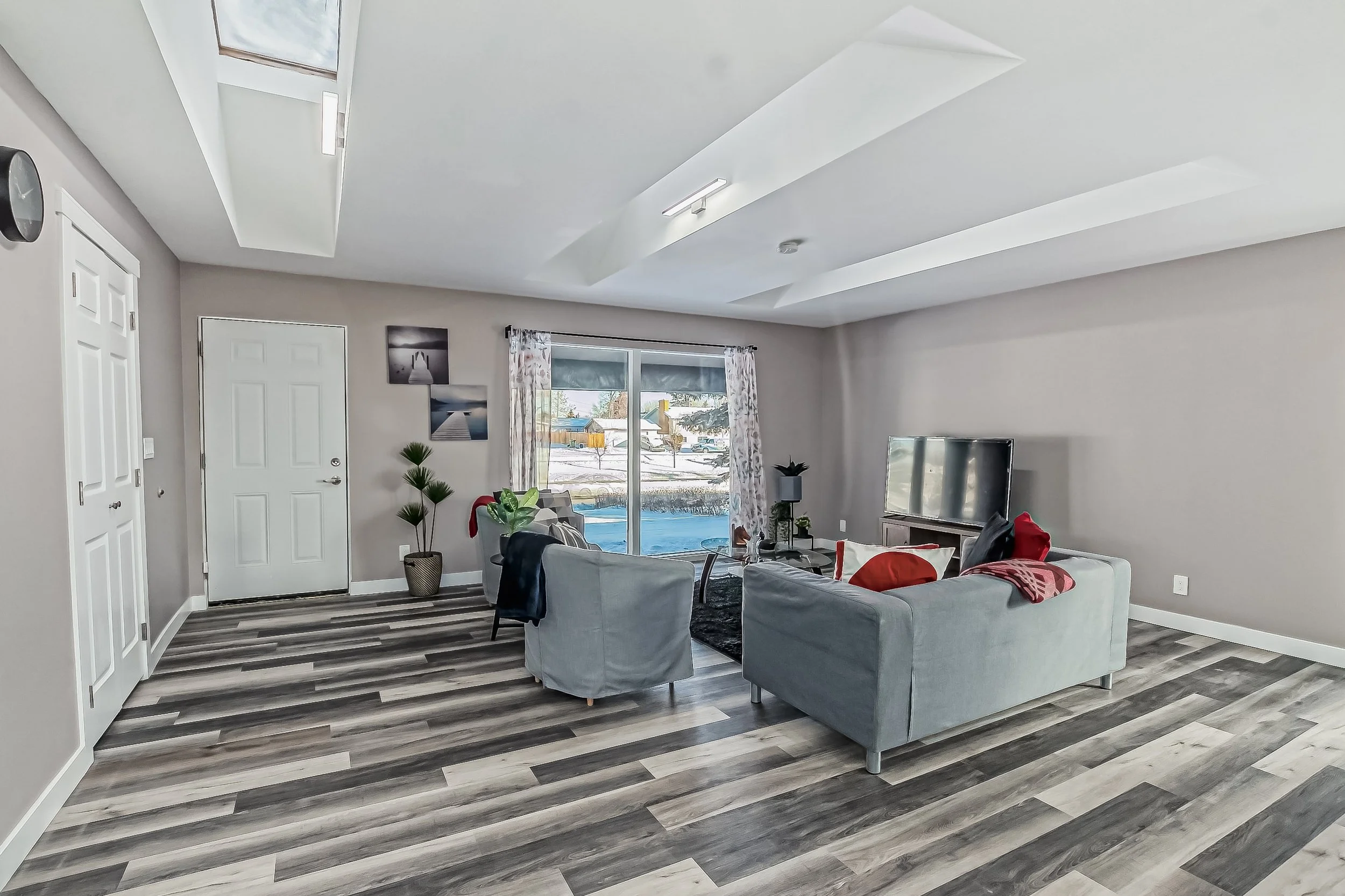 Living room with gray couches, a television on a stand, a sliding glass door with curtains, decorated with framed pictures and plants, and a view of a snowy outdoor scene.
