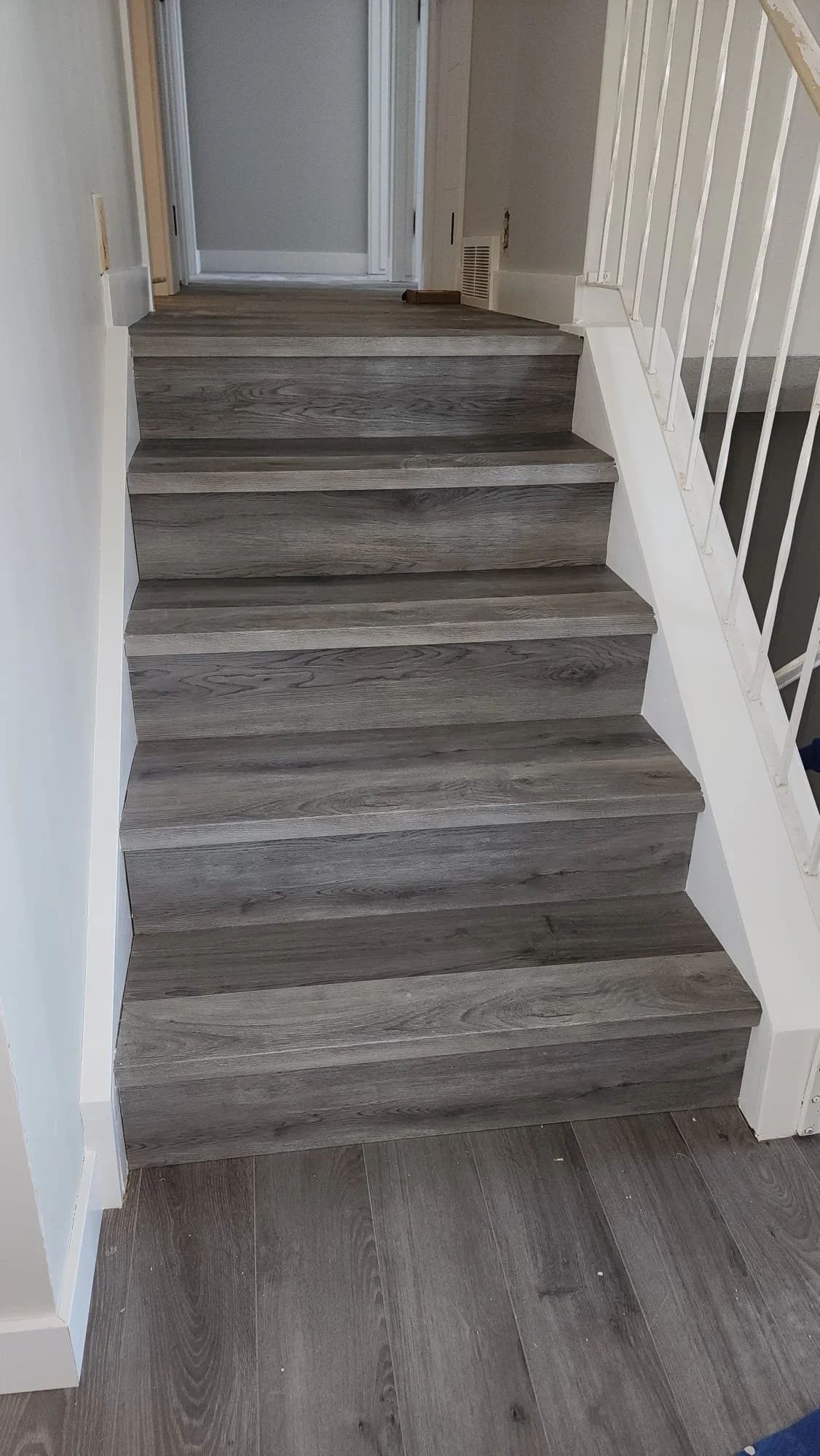 Wooden staircase with four steps leading up to a door. The steps have a grayish wood grain finish. There is a white banister on the right side and a floor with matching wood grain near the bottom of the stairs.