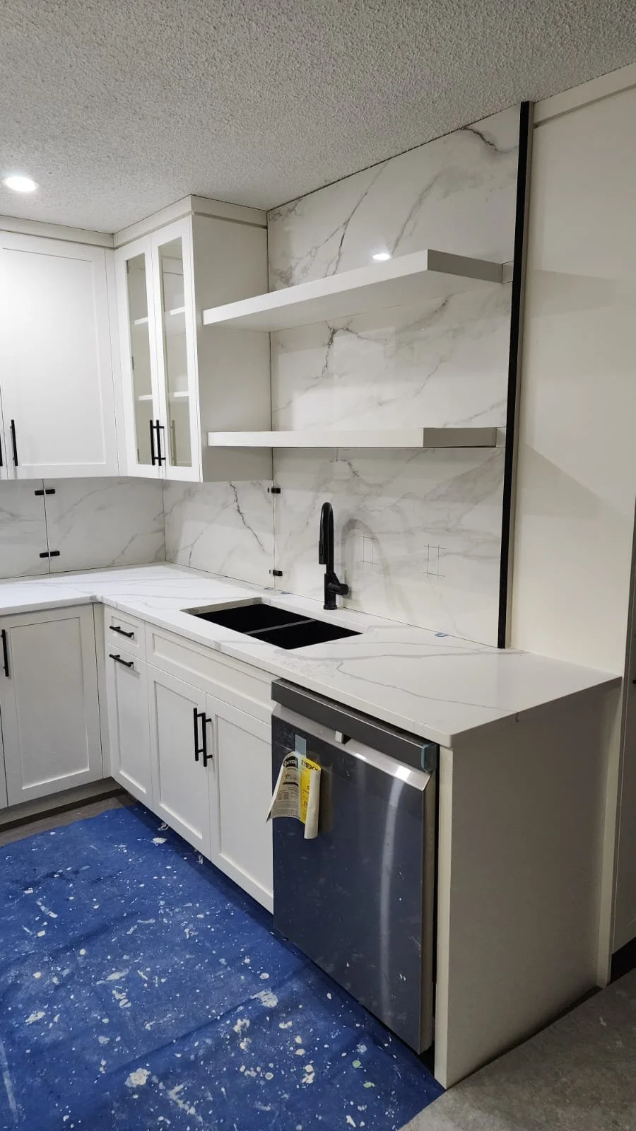 Modern kitchen with white cabinets, black hardware, marble backsplash, and black faucet, under construction with blue protective floor covering.
