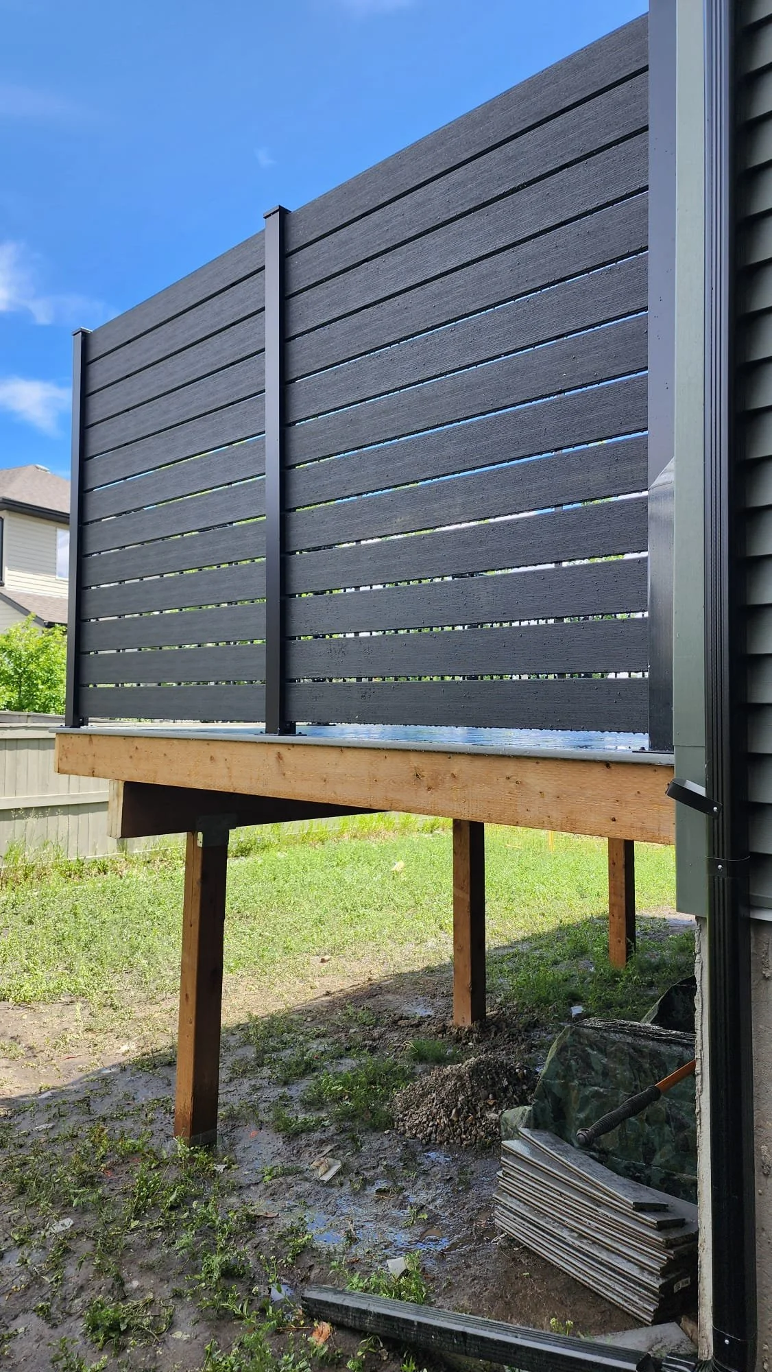 Newly built elevated deck with black horizontal siding, supported by wooden posts, adjacent to a house with dark siding, in a backyard with grass and construction materials.