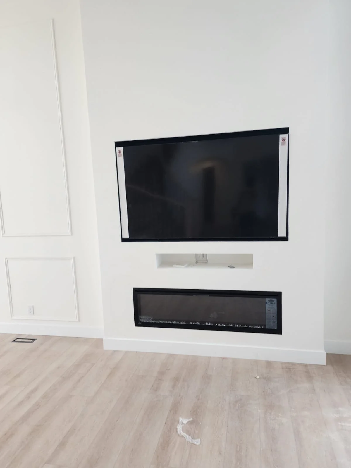 A modern living room with a mounted flat screen TV above a linear electric fireplace on a white wall, with light-colored hardwood flooring.