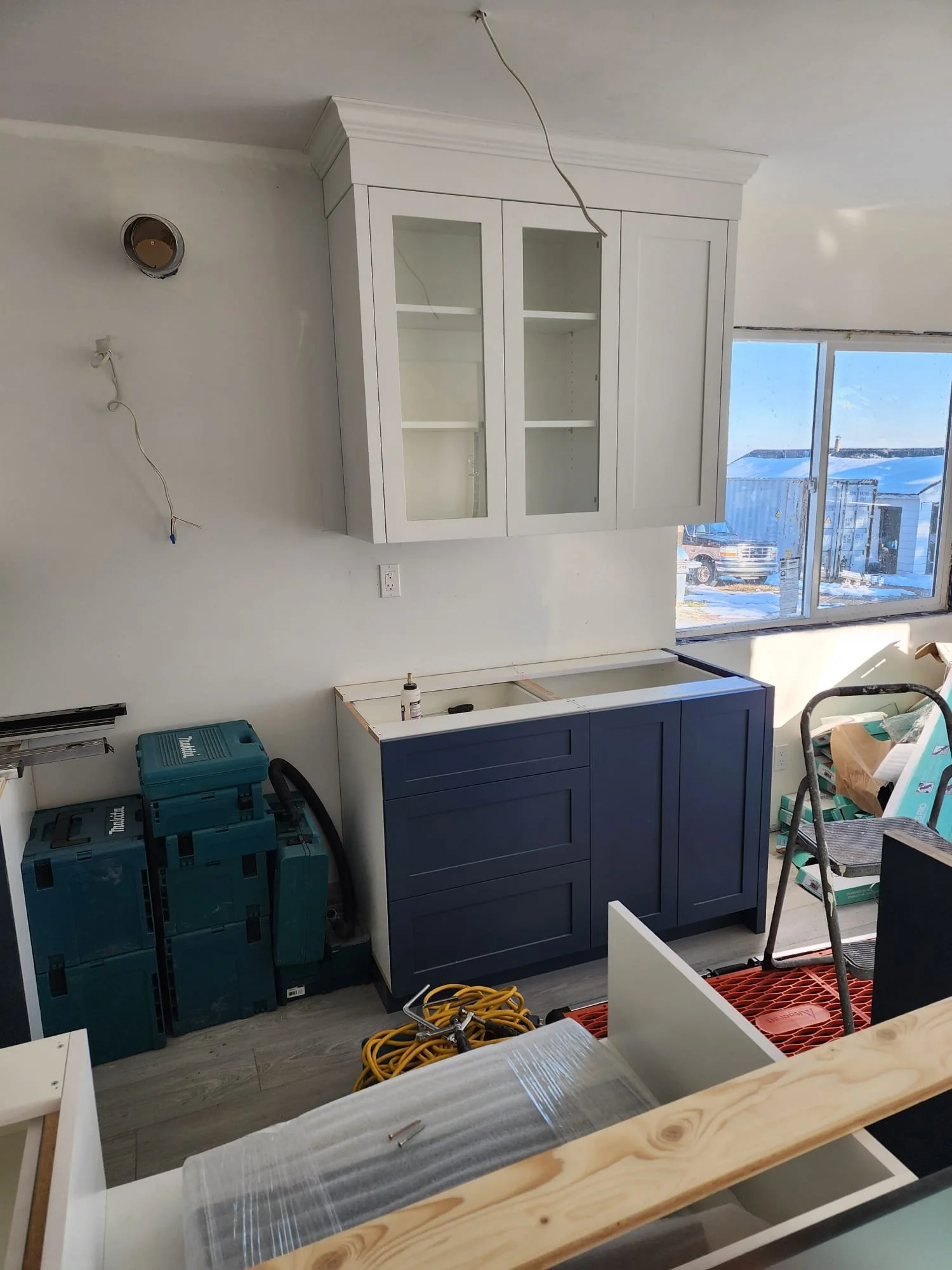 Interior of a kitchen under renovation with white wall cabinets, a dark blue base cabinet, construction tools, and a window with a view of a snowy landscape.