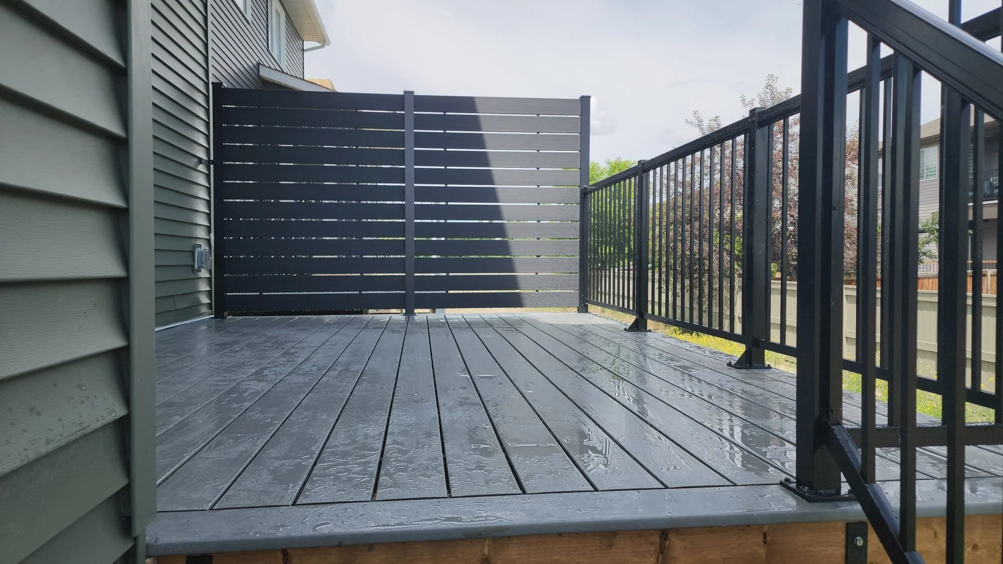A newly installed wooden deck with black metal railing on a residential house, wet from recent rain, with a privacy screen at the back and neighboring houses visible in the background.