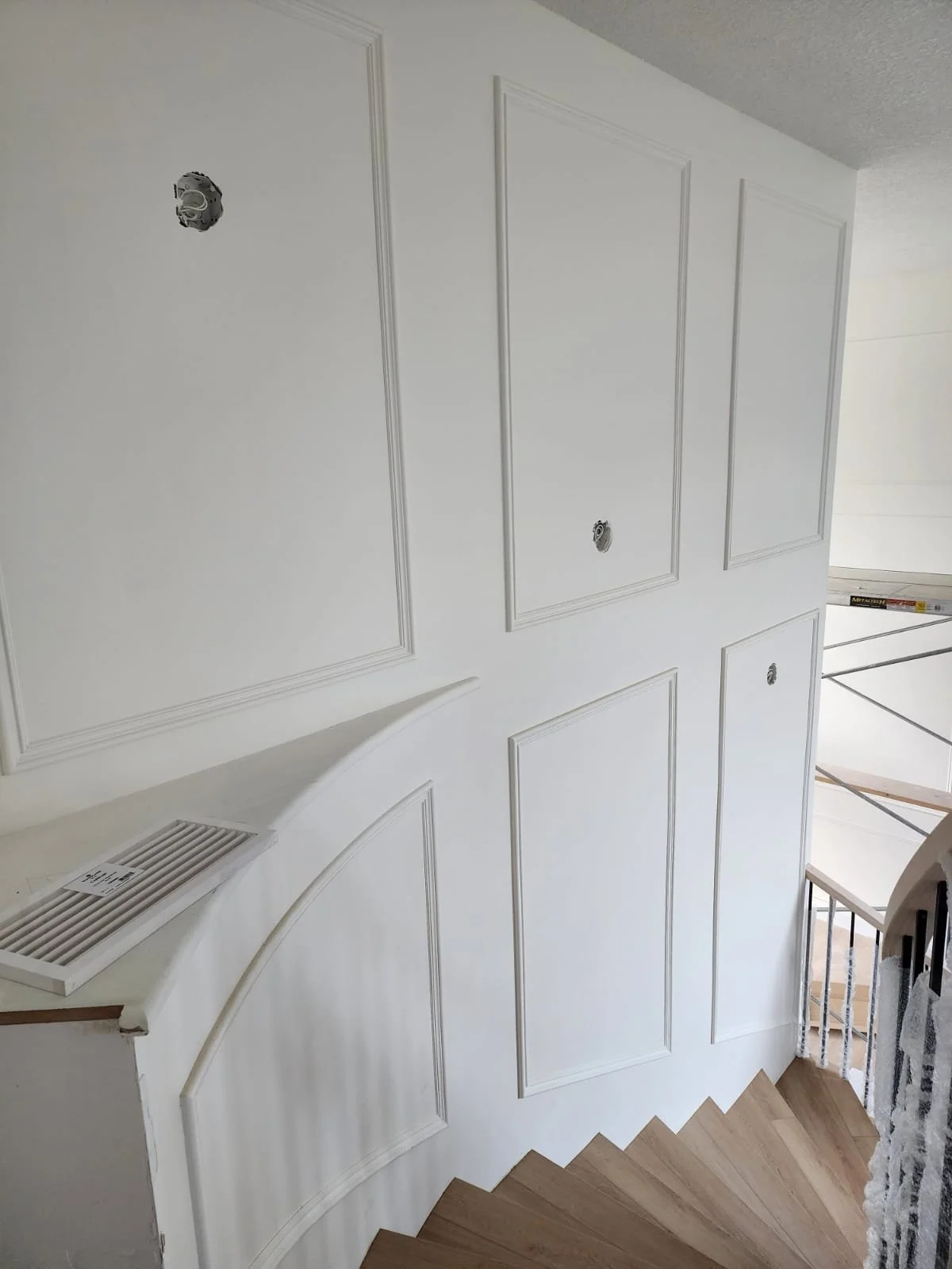 White wall with decorative molding panels, unfinished electrical boxes, partial view of staircase, and some construction materials.