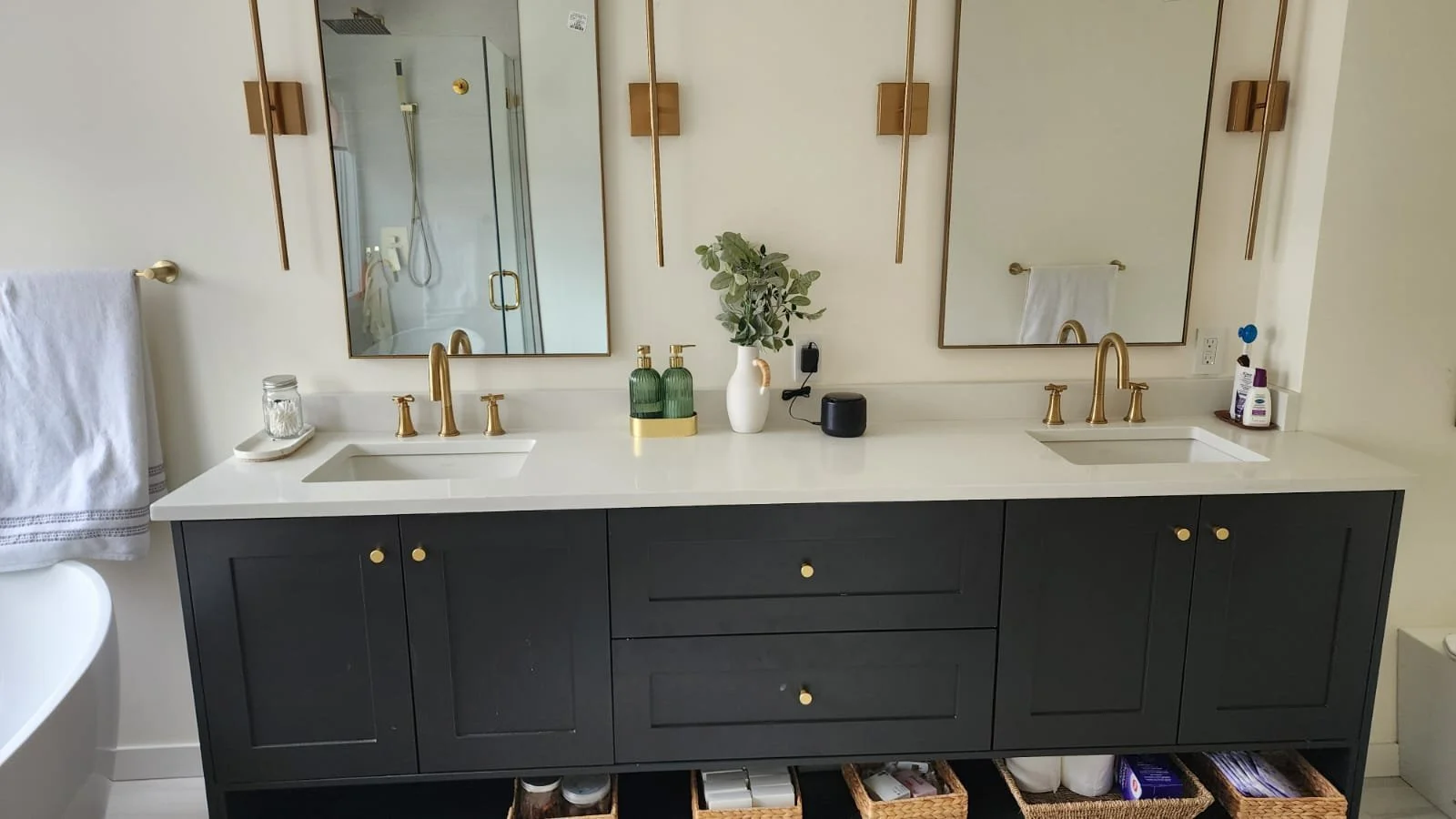 Double vanity bathroom with black cabinet, white countertop, two sinks with gold fixtures, two mirrors, wall-mounted lights, small plants, toiletries, and storage baskets underneath.