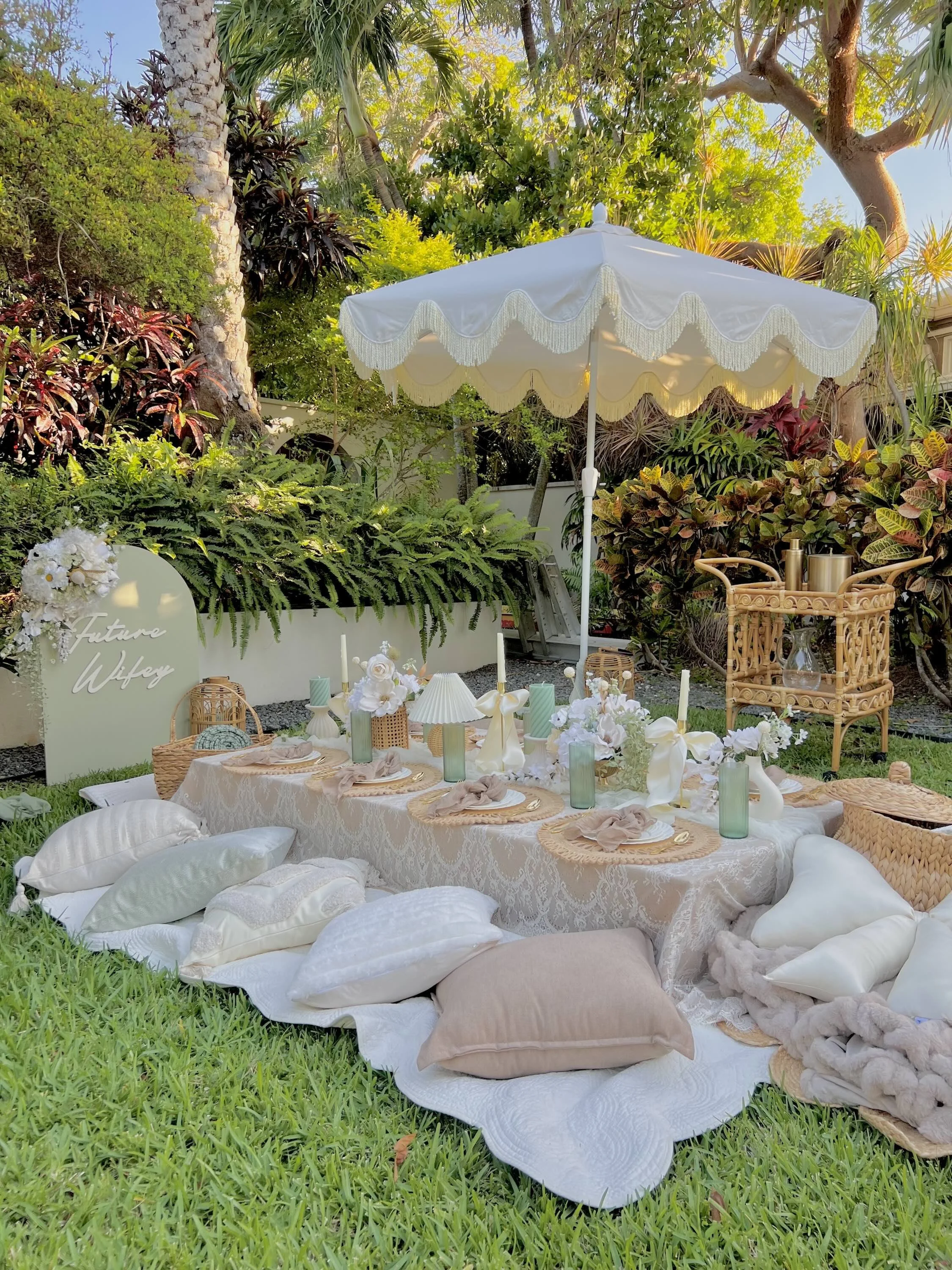 Outdoor garden setup for a wedding or special event with a low table covered with a white lace tablecloth, pillows arranged around it, and decor including flowers, candles, and tableware under a white umbrella with fringe. There is a sign that reads 'Future Wife' and lush greenery in the background.