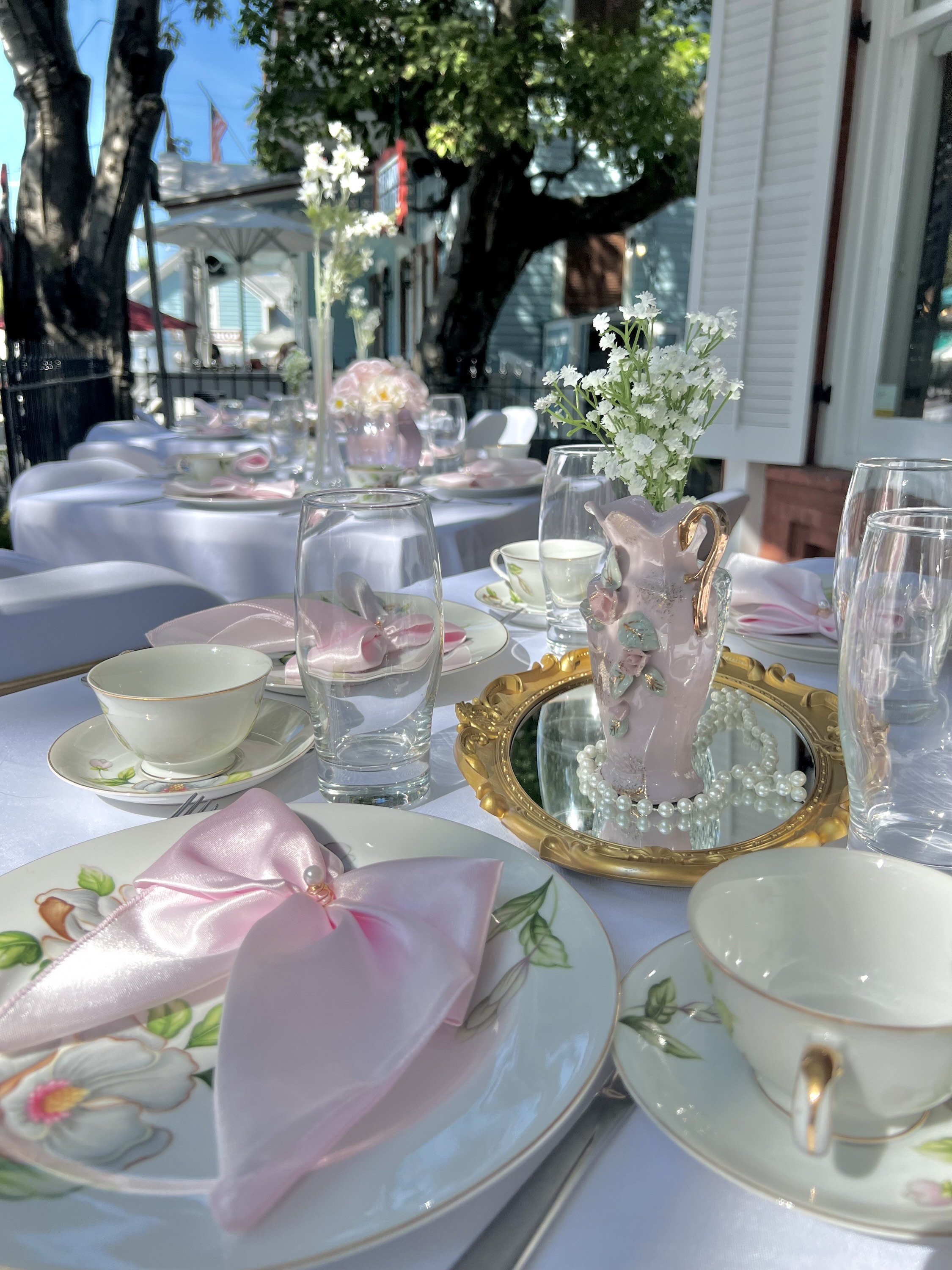 Elegant outdoor table set for afternoon tea with floral china, pink napkins, glassware, and a centerpiece of white flowers in a pink vase with pearl accents, under trees with a house and fence in the background.