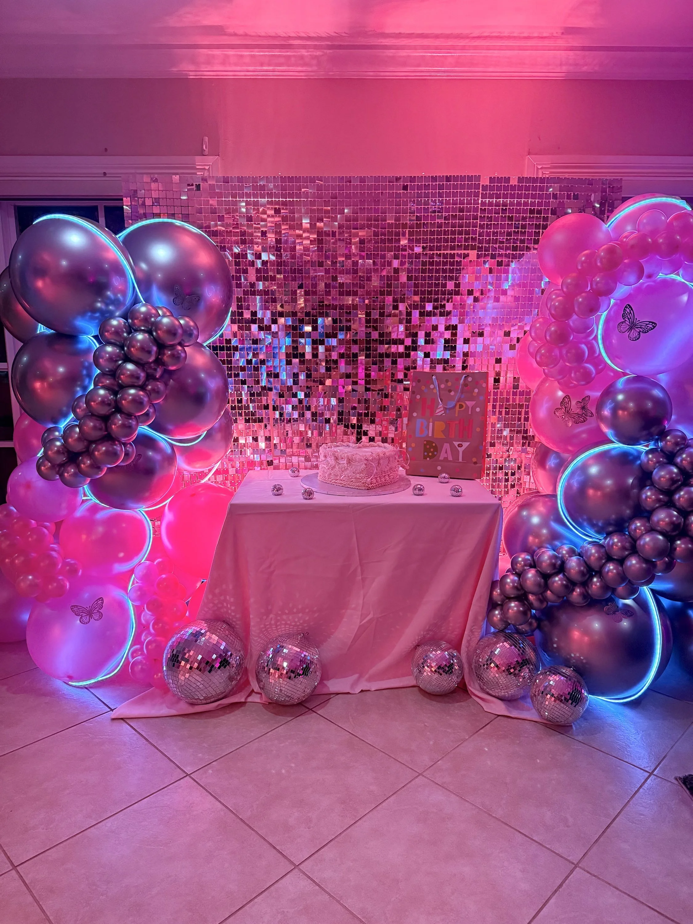 Pink-themed birthday party setup with a cake on a table, surrounded by pink and silver balloons, disco balls, and a holographic backdrop with a colorful 'Happy Birthday' sign.