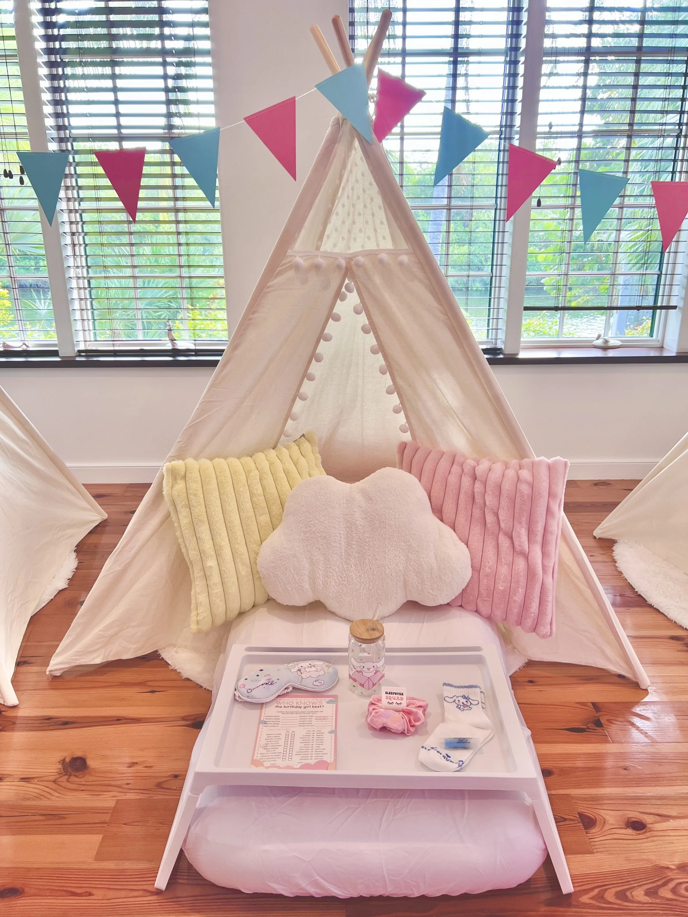 Decorative children's tipi tent with pink and blue bunting, two plush pillows (yellow and pink), and a cloud-shaped pillow, set up indoors with a wooden floor and large windows.