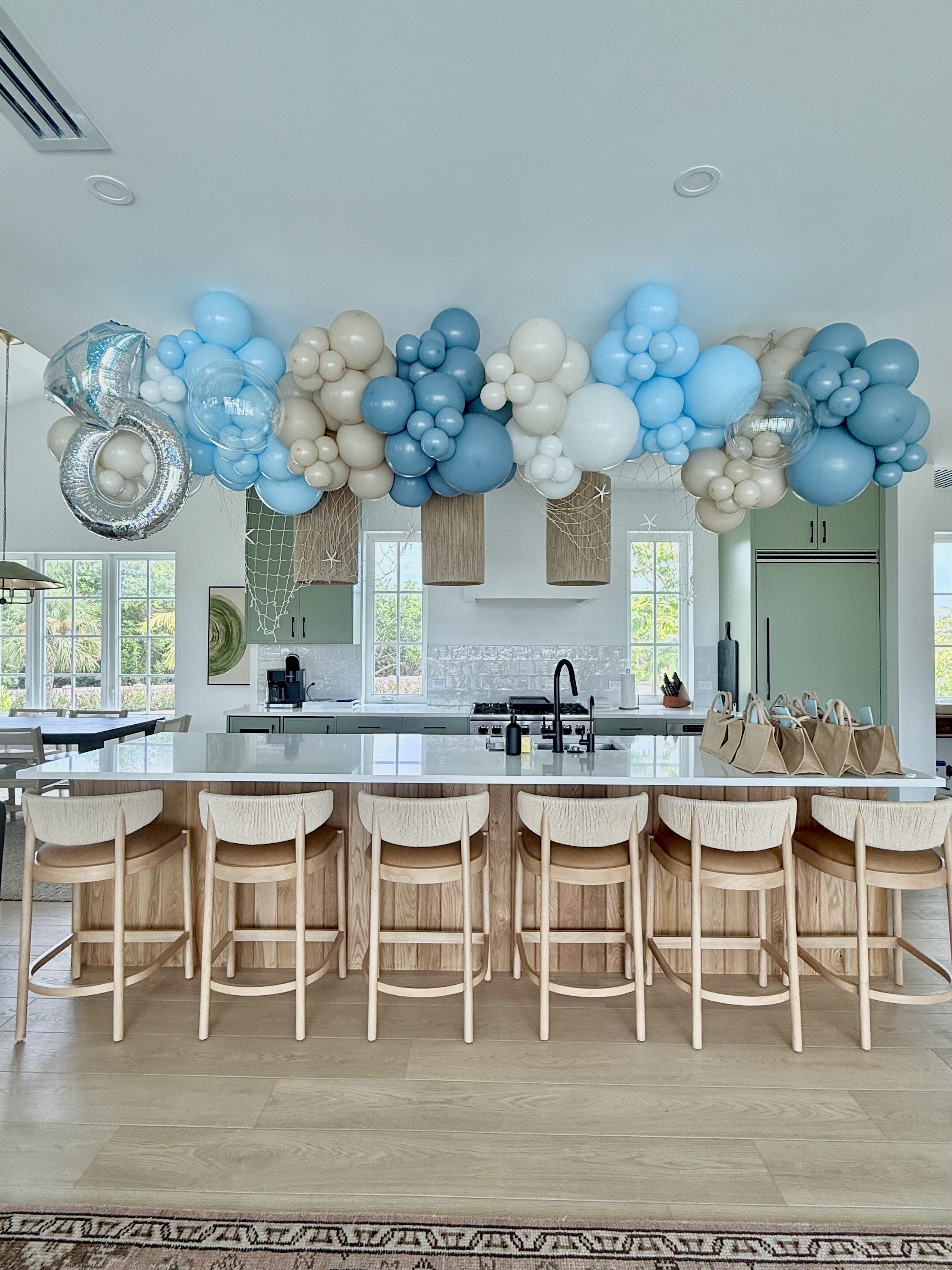 Decorative balloon arrangement in shades of blue, white, and beige hanging above a kitchen island with beige bar stools.