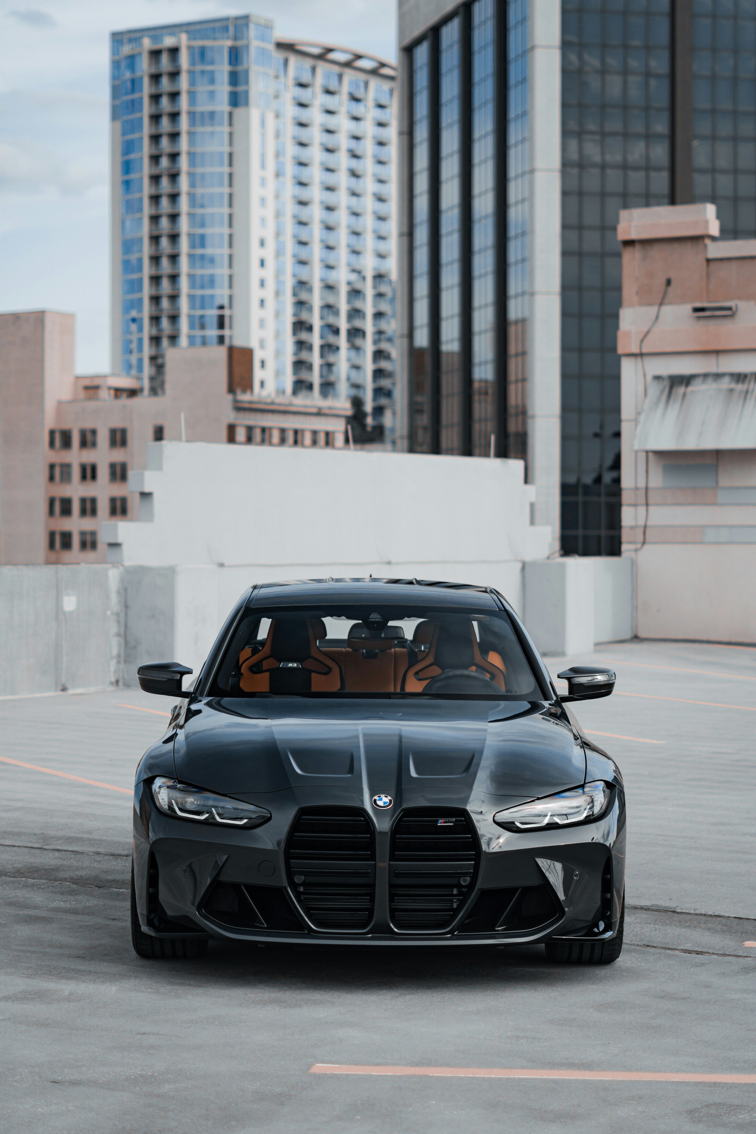 A black BMW M Series sports car parked on the top level of a parking garage in an urban setting with tall modern office buildings in the background.