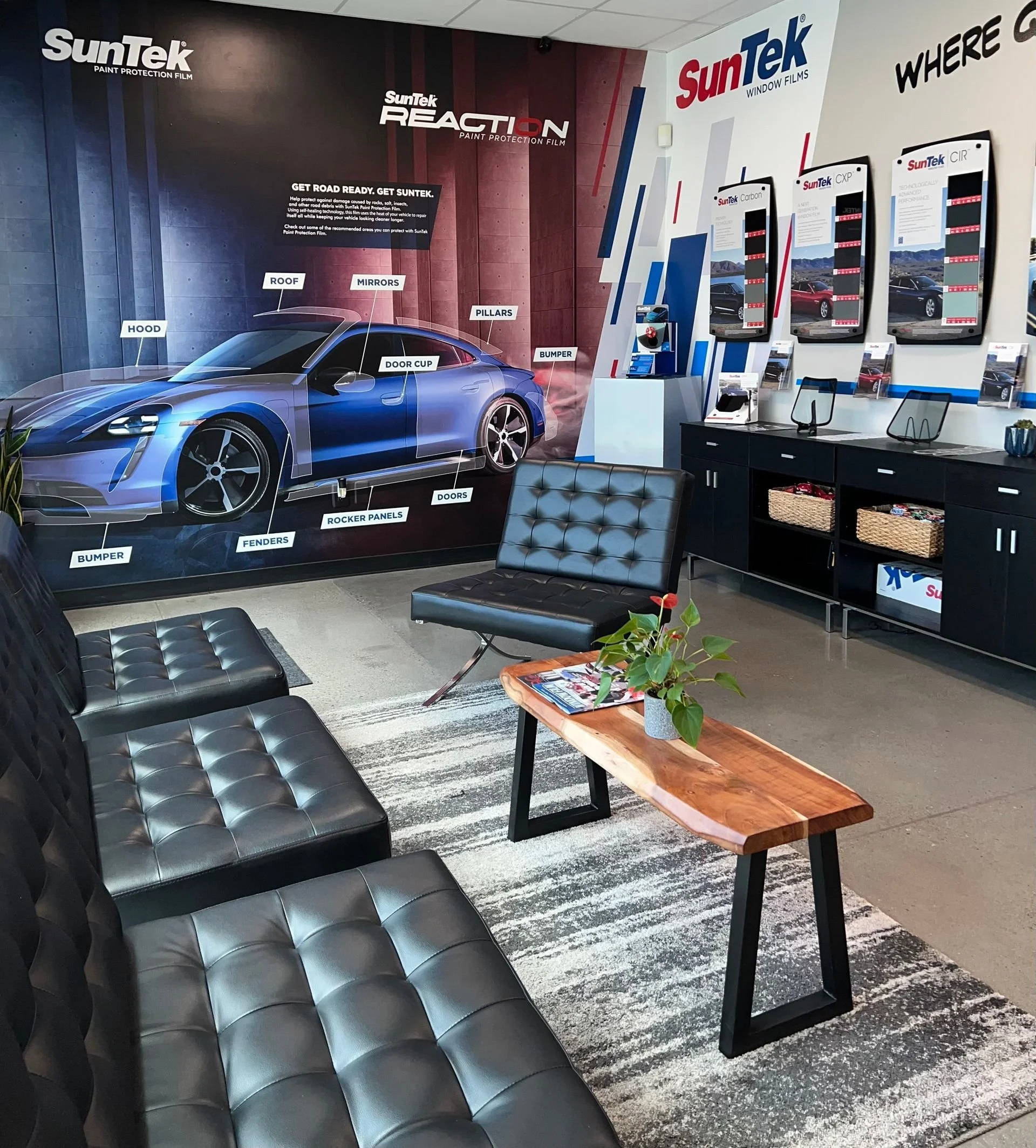 Interior of a SunTek window film showroom with a large illustrative wall poster of a sports car labeled with branding and car parts, a seating area with black leather chairs, a wooden coffee table with a plant, and a black cabinet with samples and brochures displayed.