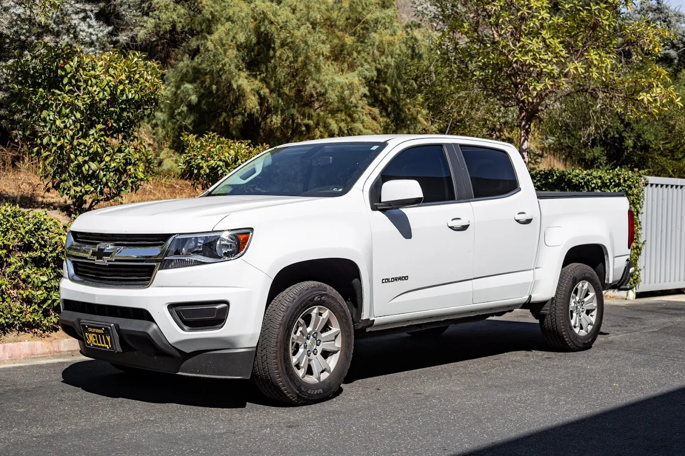 White Chevrolet Colorado pickup truck parked on the side of a road with green bushes and trees in the background.