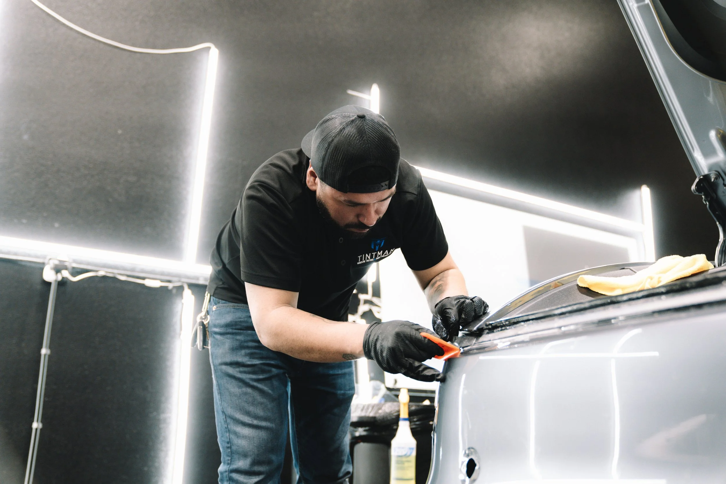A man with a black cap and black gloves shaving the edge of a silver car trunk with a small razor or scraper in a brightly lit garage.