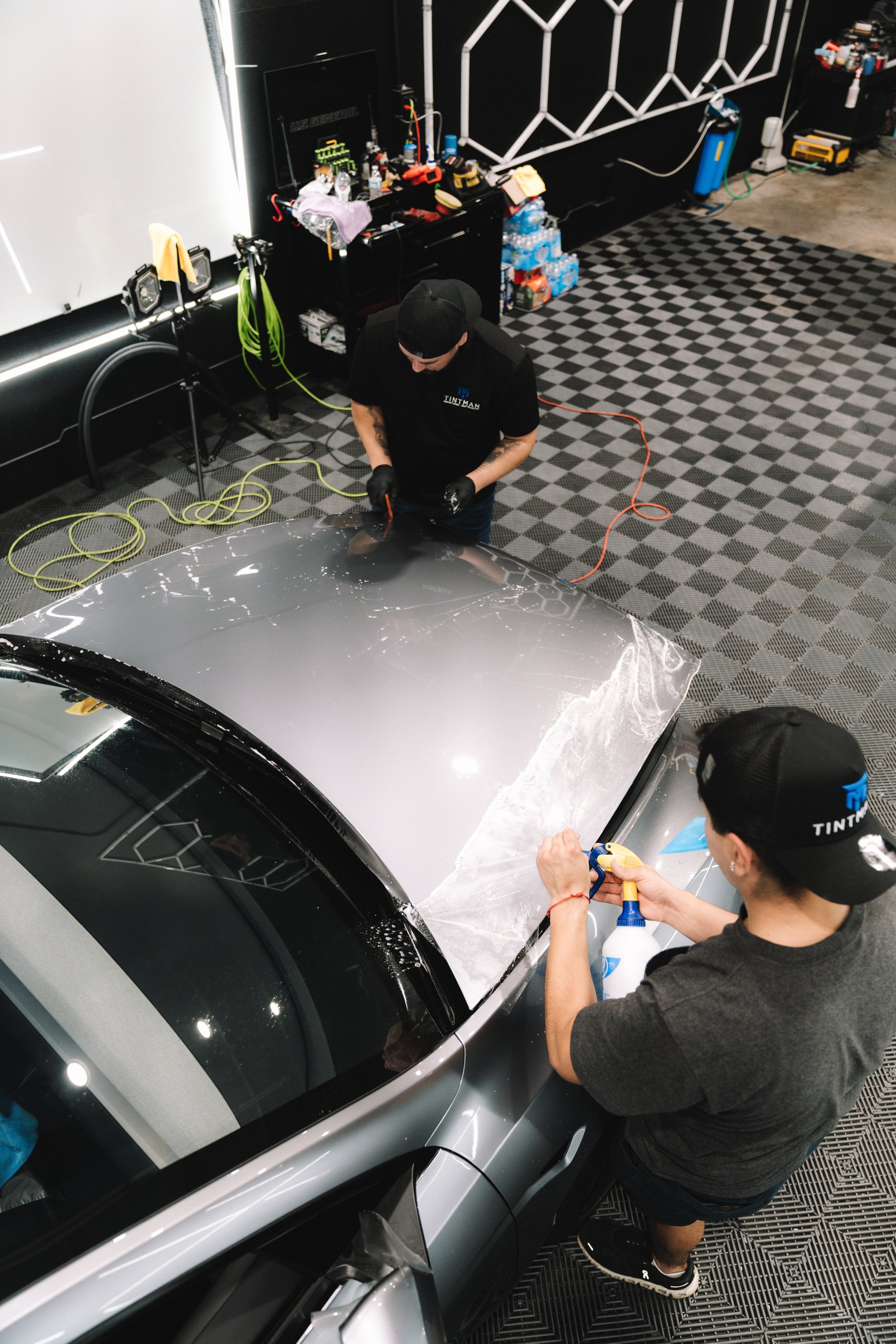 Two workers applying protective film on the hood of a silver sports car in a workshop.