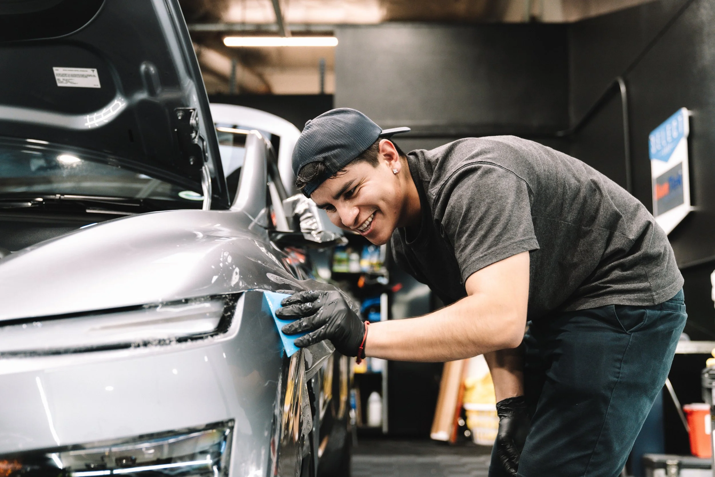 A young man wearing a black cap, grey t-shirt, and black gloves polishing a silver car in an auto repair shop.