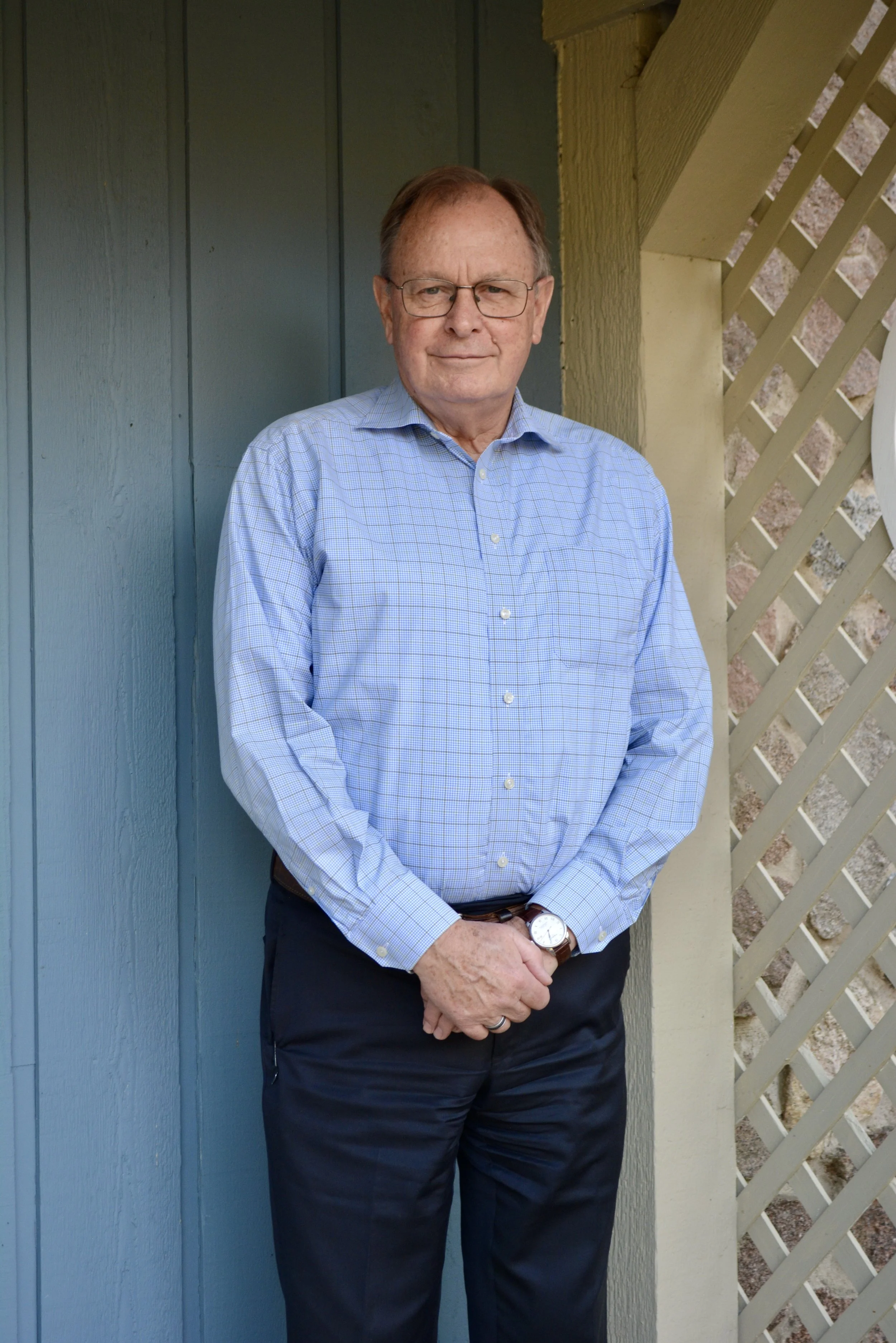 An elderly man with glasses wearing a light blue checkered shirt and dark pants standing outdoors next to a blue wooden wall and beige lattice fence.