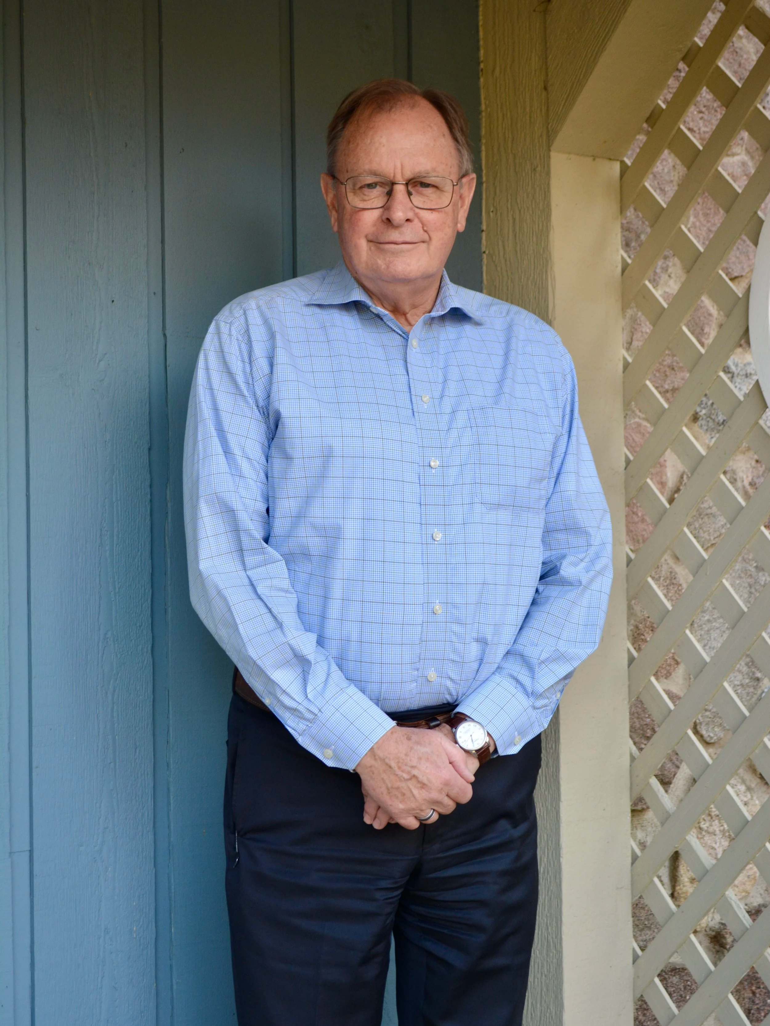 A senior man with glasses, wearing a light blue checkered shirt and dark pants, standing outdoors near a blue wooden wall and a lattice fence.