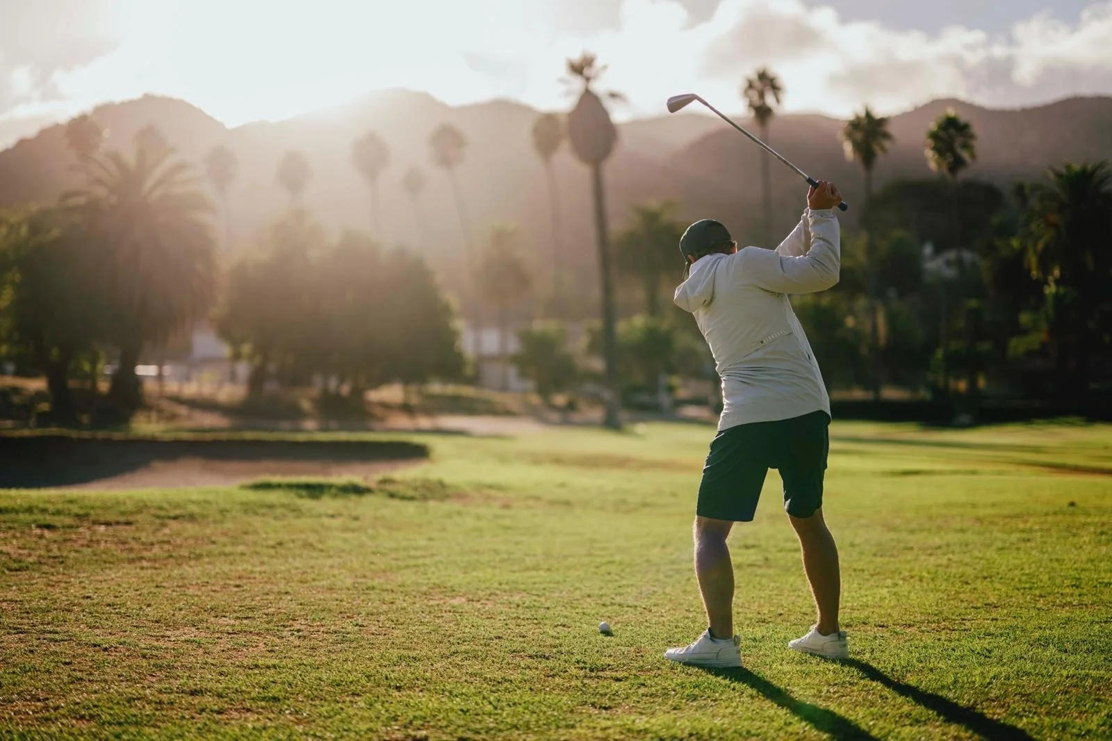 A man playing golf on a sunny course with palm trees and mountains in the background.