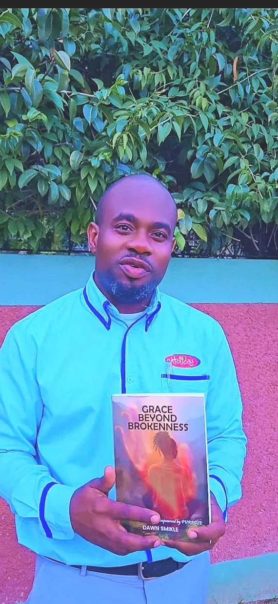 A man in a light blue shirt holding a book titled "Grace Beyond Brokenness" by Dawn Smikle. He stands outdoors in front of a green leafy background with a wall painted in blue and red.