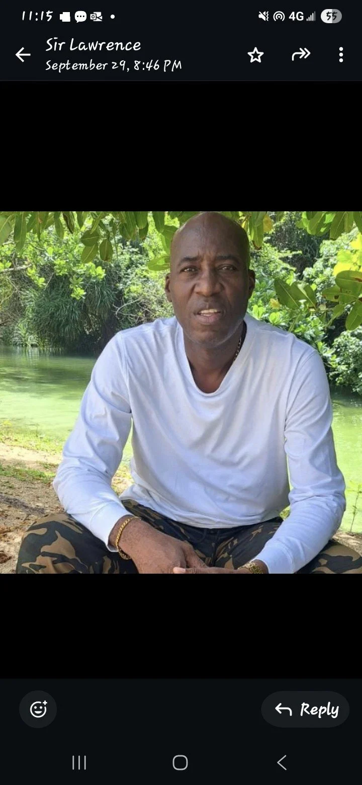 A man sitting outdoors near a body of water surrounded by lush green trees, wearing a white long-sleeve shirt and camouflage pants.