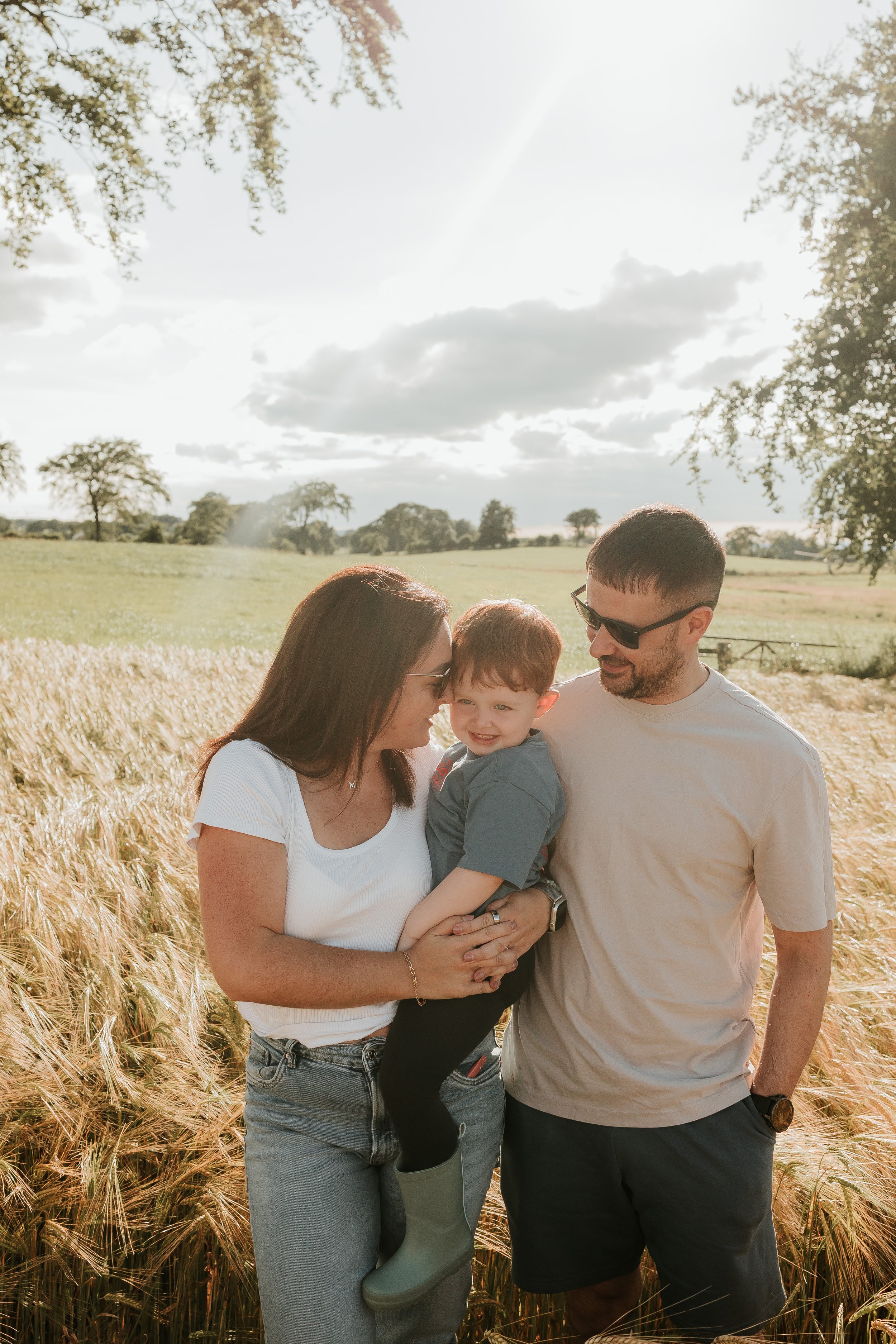 A family of three, a woman, a man, and a young boy, standing in a wheat field on a sunny day, smiling and talking.