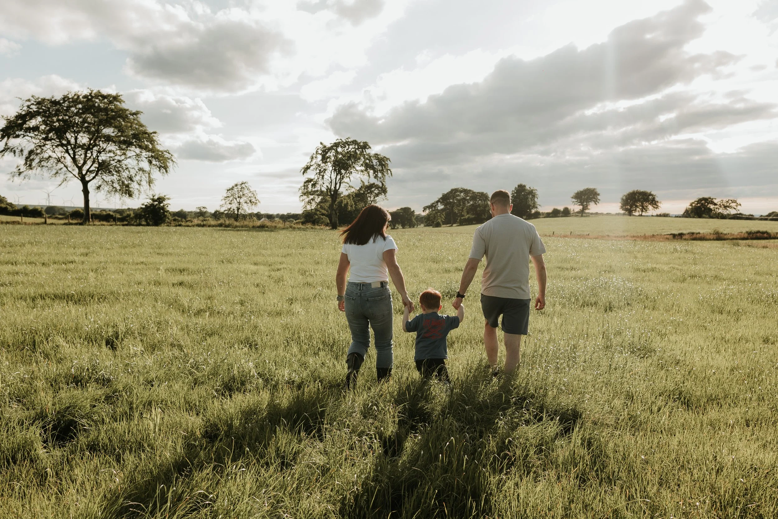 A family of three walking hand in hand through a grassy field on a cloudy day.