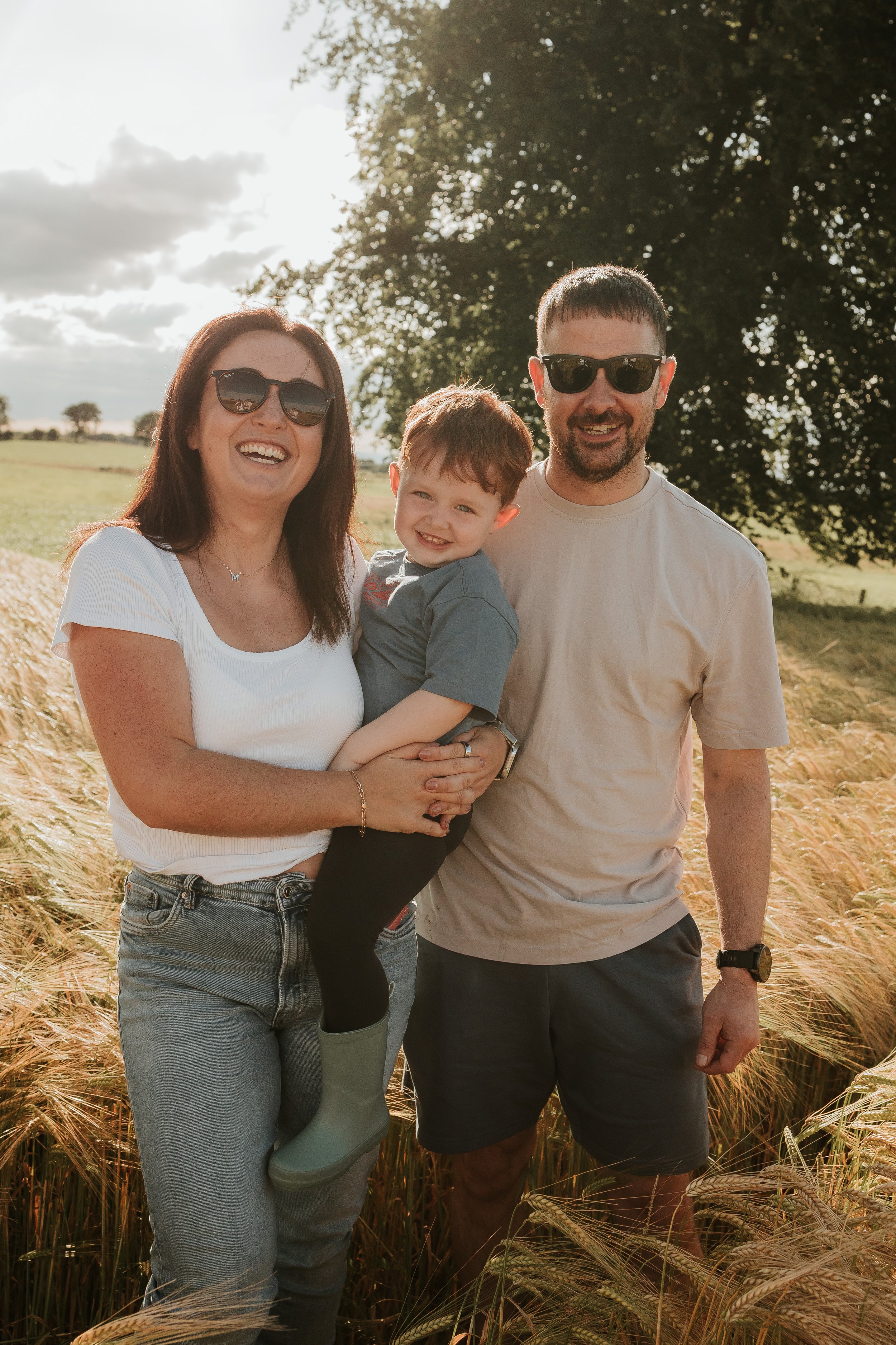 A family of three standing outdoors in a wheat field with trees in the background, smiling and looking at the camera.