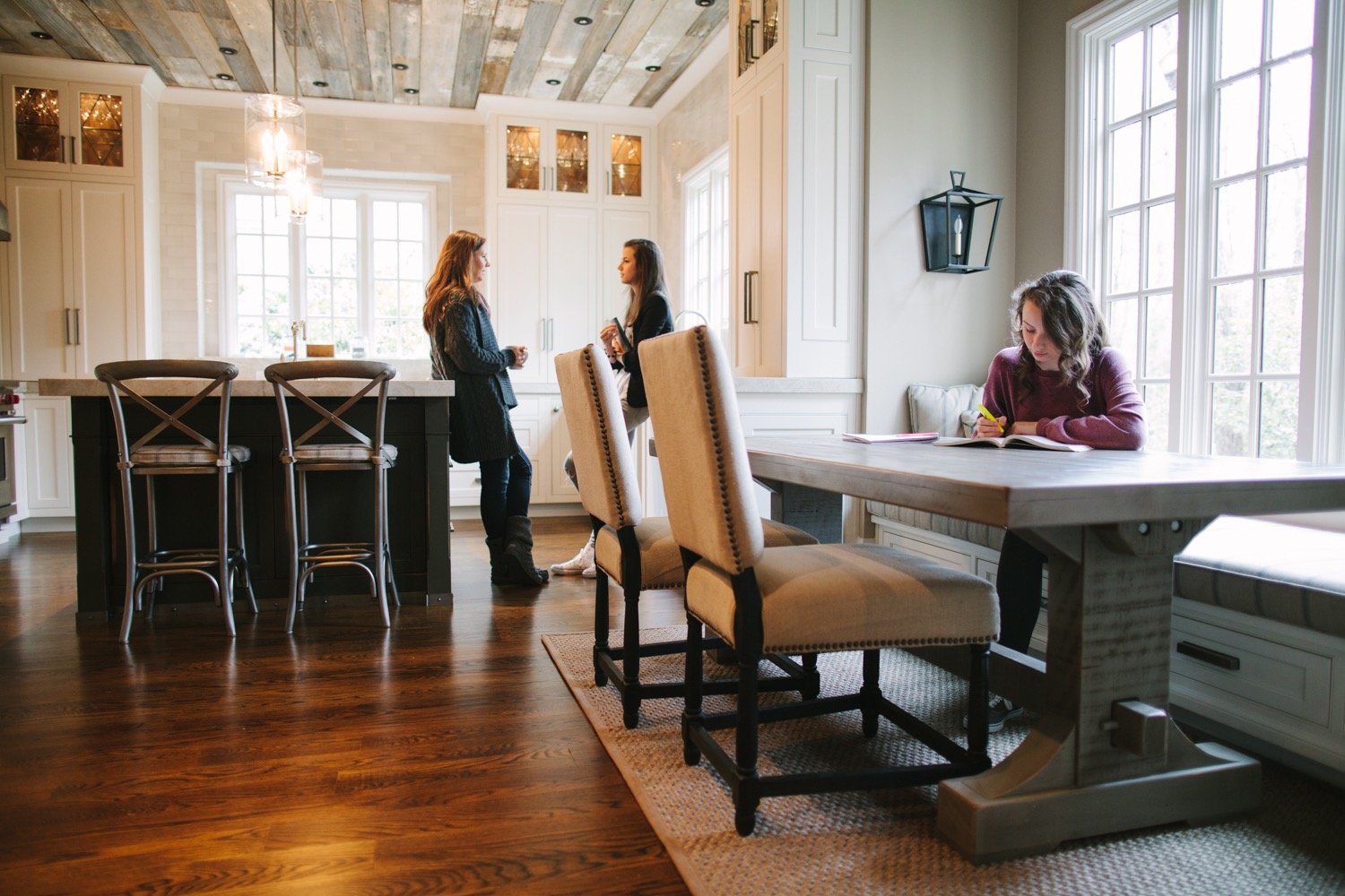 A spacious kitchen with a dining table, chairs, and a kitchen island. Two women are standing and talking near a large window, one is holding a coffee mug, the other has a phone. A woman is sitting at the table, writing in a notebook.