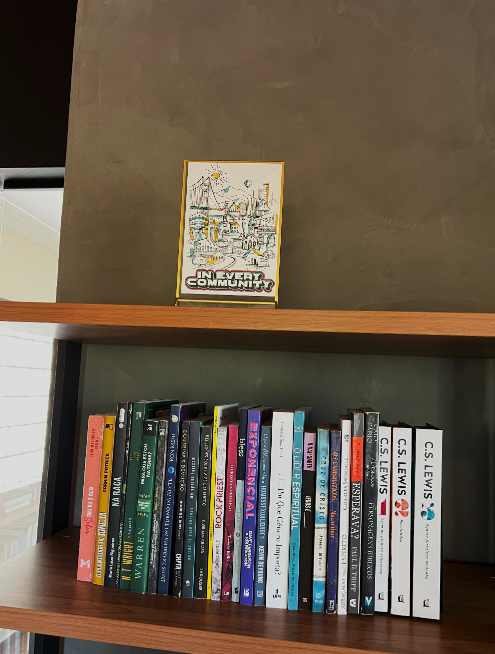 A bookshelf with various books on society, culture, and spirituality, with a sign above that reads 'In Every Community' featuring a colorful illustration of a cityscape.