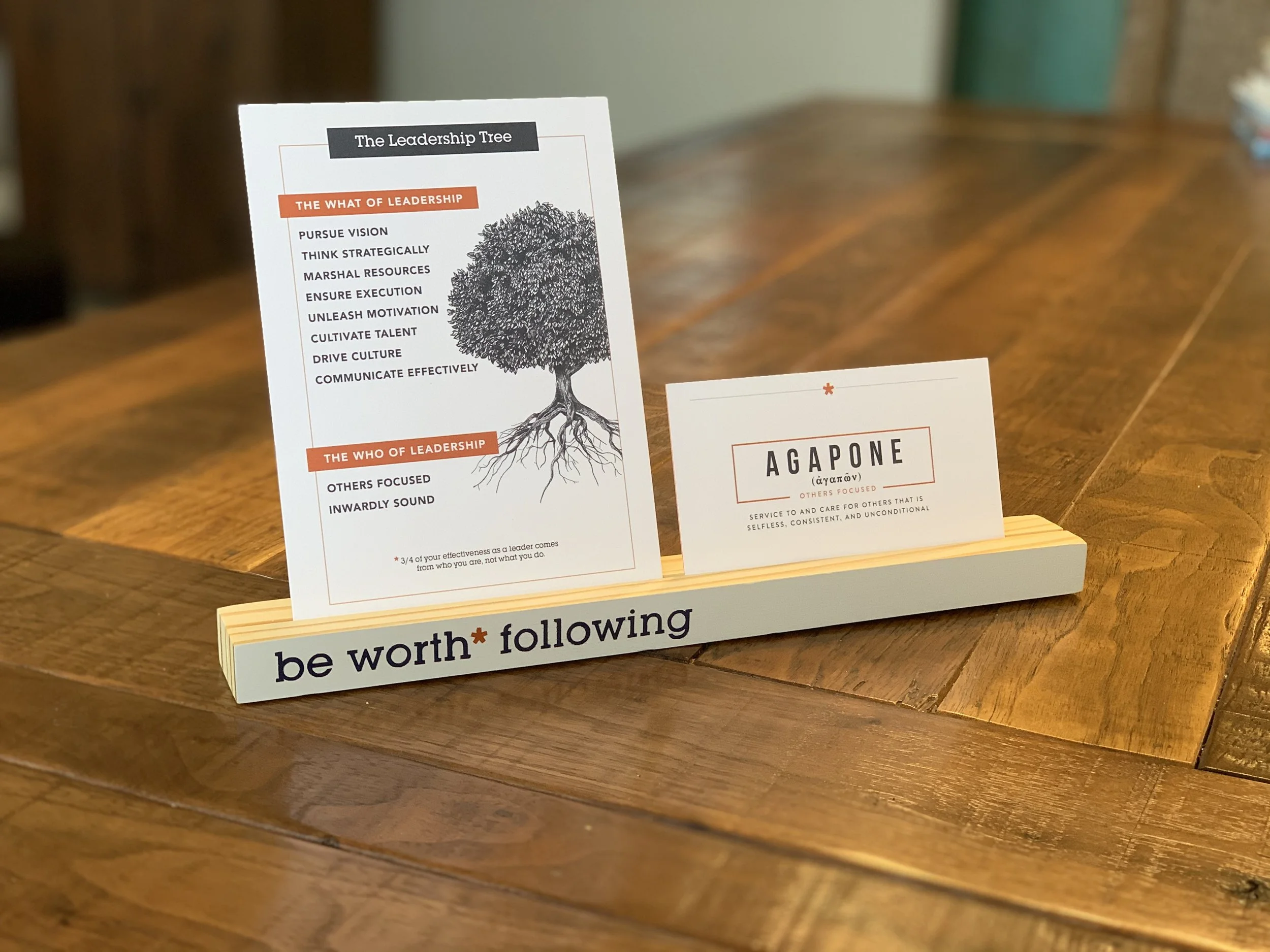 A small desk sign displays leadership principles and a card with the word Agapone, emphasizing service and selflessness, on a wooden table.
