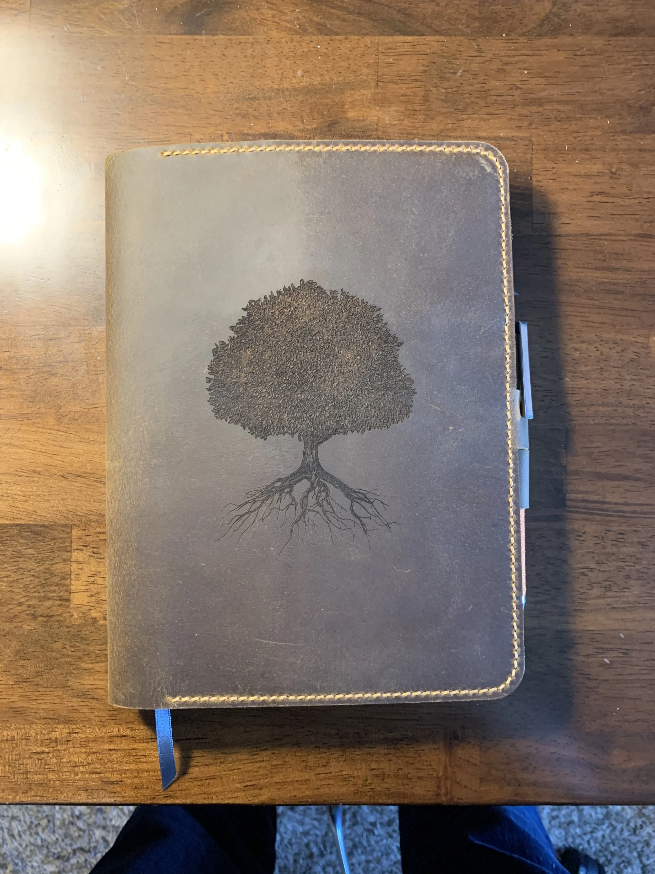 A closed brown notebook with a black tree and roots illustration on the cover, placed on a wooden surface.