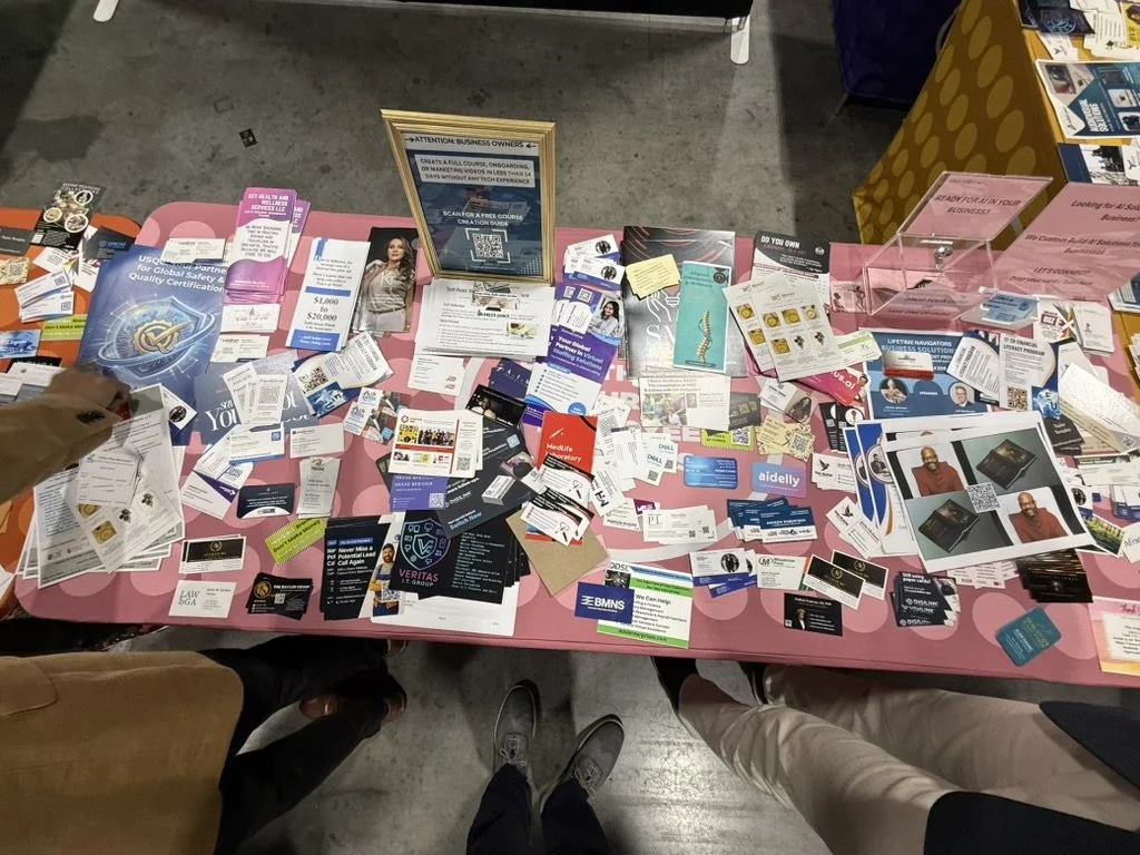 A pink table covered with business cards, flyers, brochures, and promotional materials at a convention or trade show.