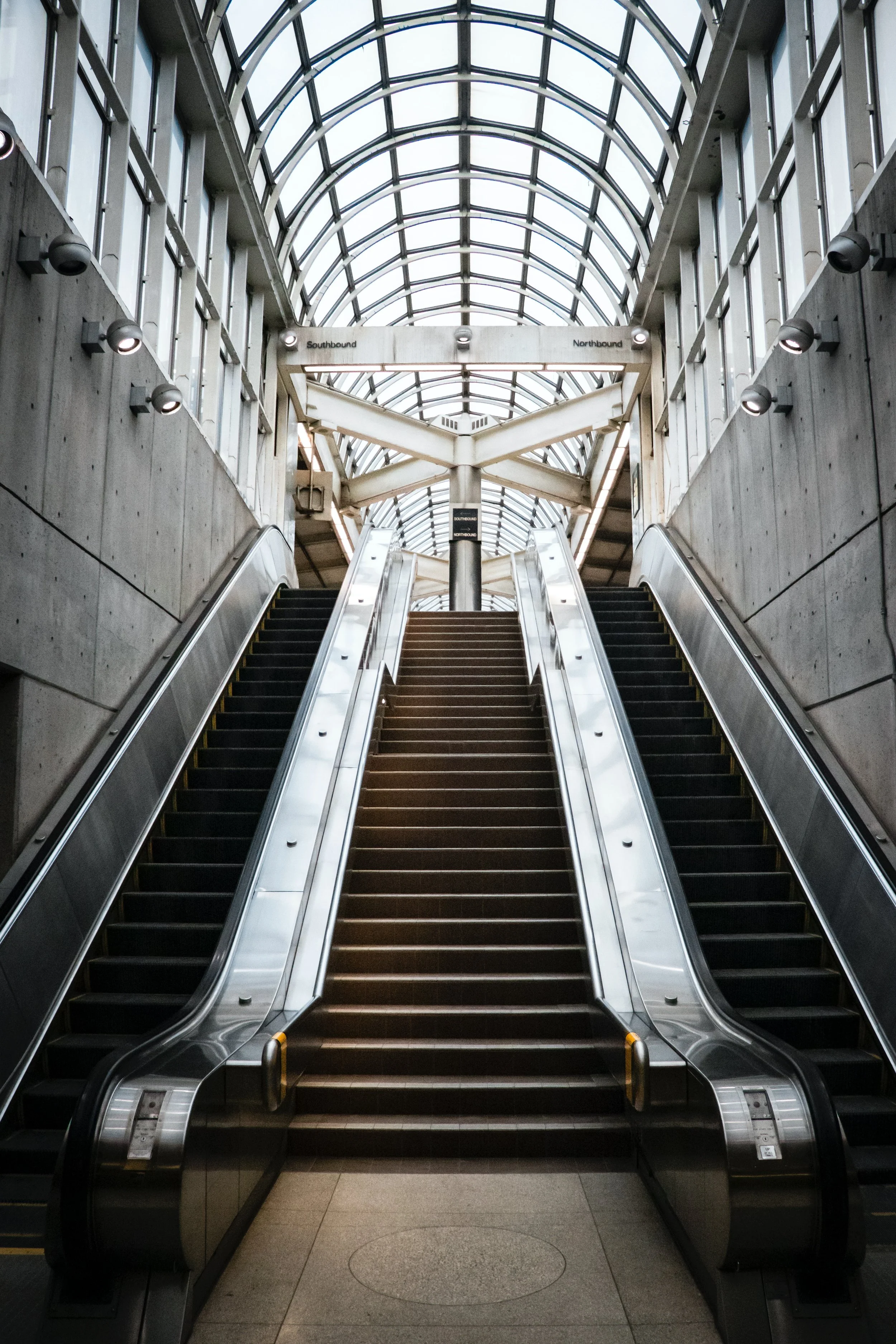 Indoor escalators leading up in a modern, glass-roofed building with concrete walls.