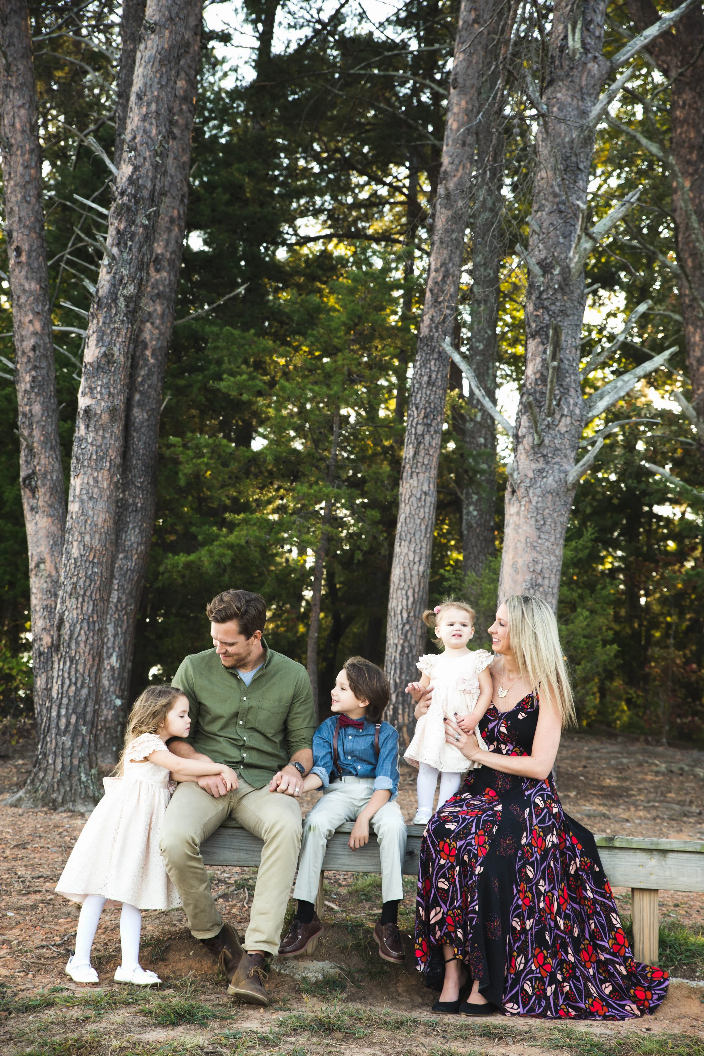 A family of five, including two women and three children, sitting on a wooden bench in a forested area with tall trees, during daylight.