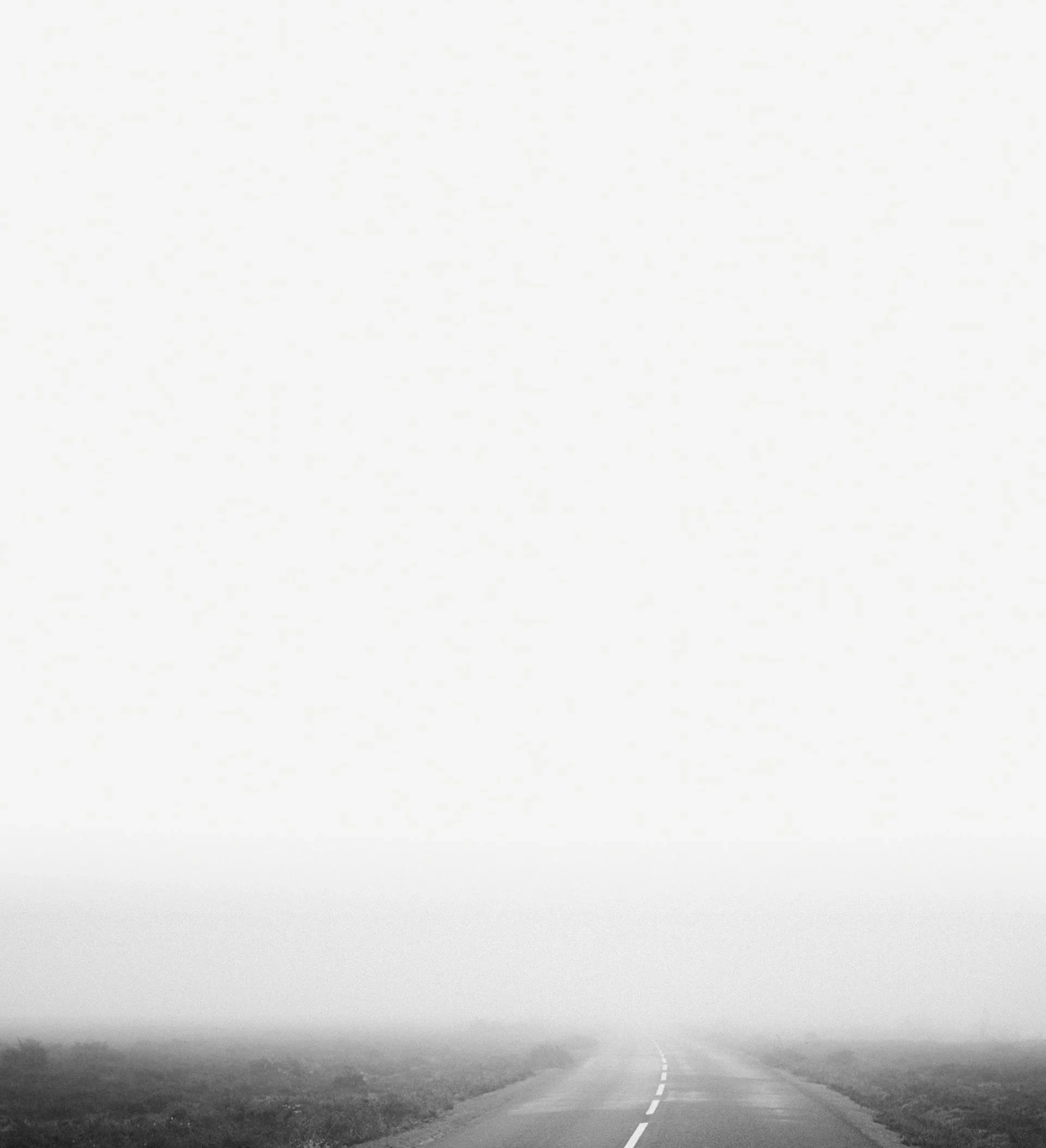 A black and white photo of an empty rural road stretching into the foggy distance with flat land on either side.