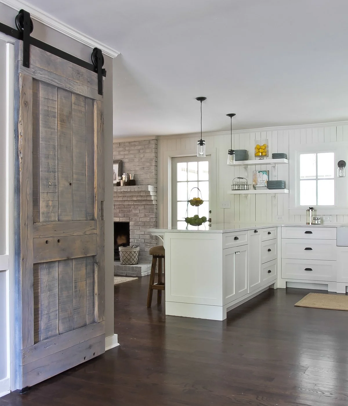 Modern kitchen with white cabinets, dark wood flooring, open shelves with dishes and yellow decorative items, pendant lighting, and a barn-style sliding door.