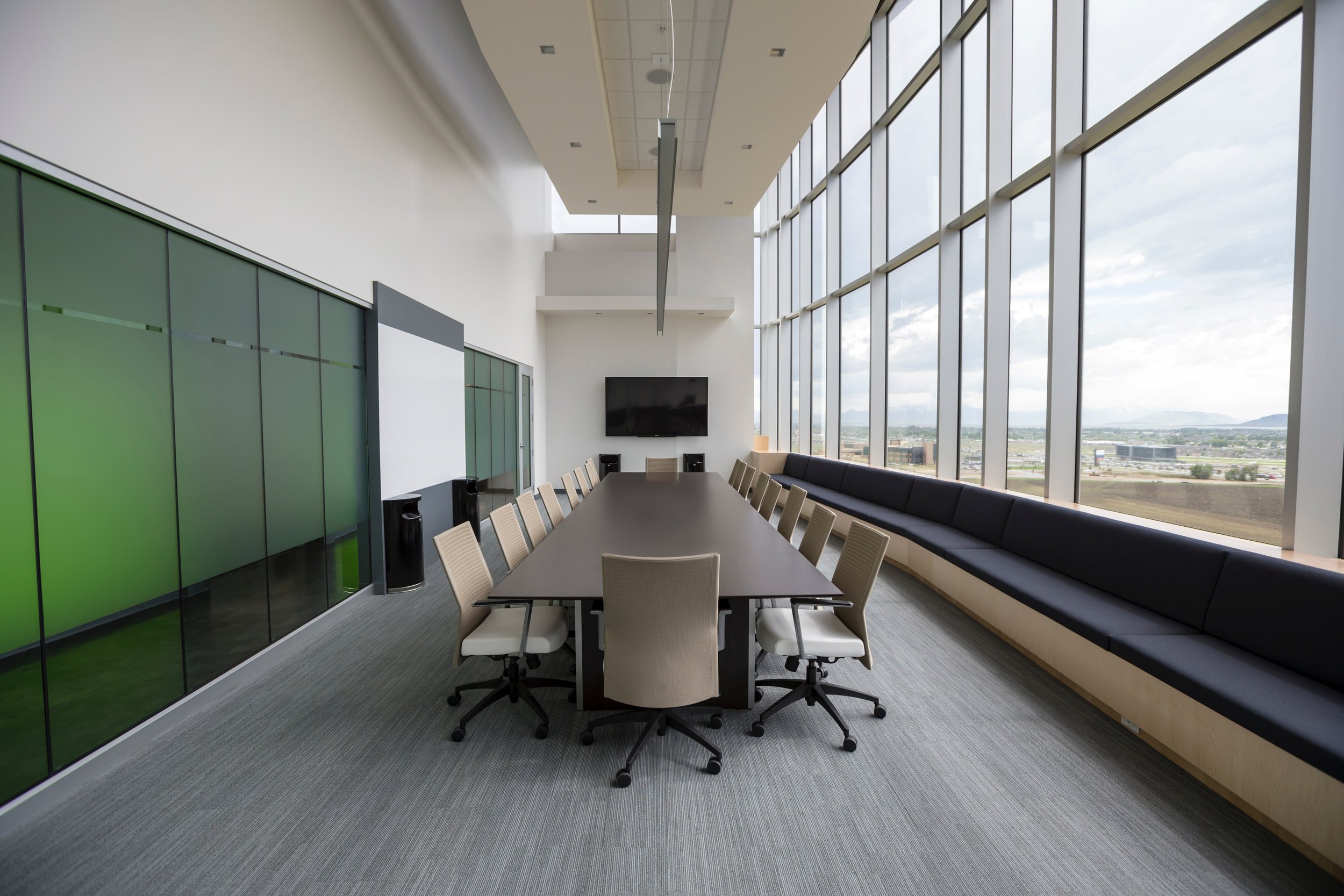 Modern conference room with a long rectangular table surrounded by white swivel chairs, large windows with a city view, a wall-mounted television, and a built-in bench along the windows.